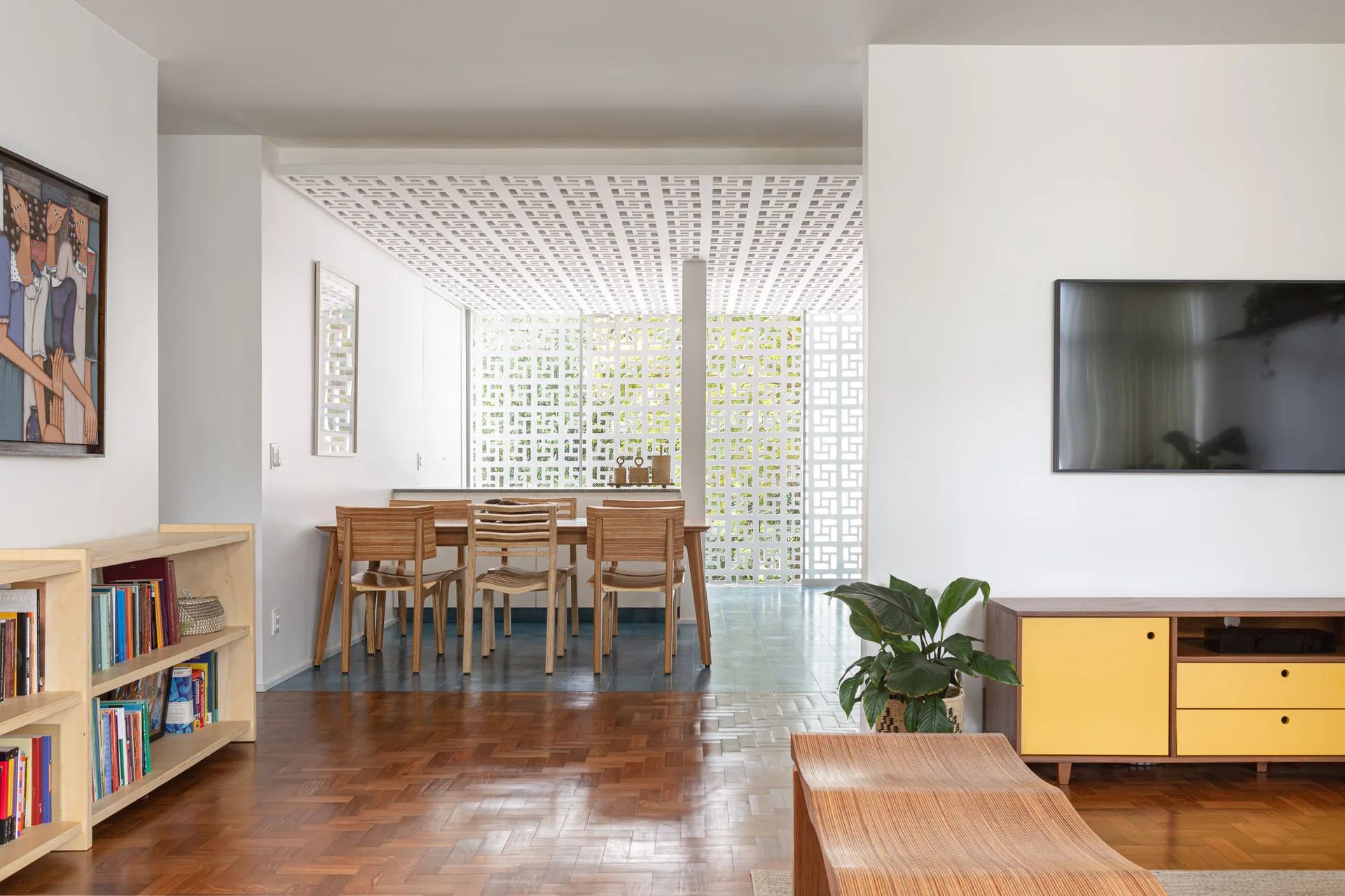 An open-plan living and dining room with a wooden dining set, parquet flooring, yellow media console, and a geometric breeze block ceiling partition.