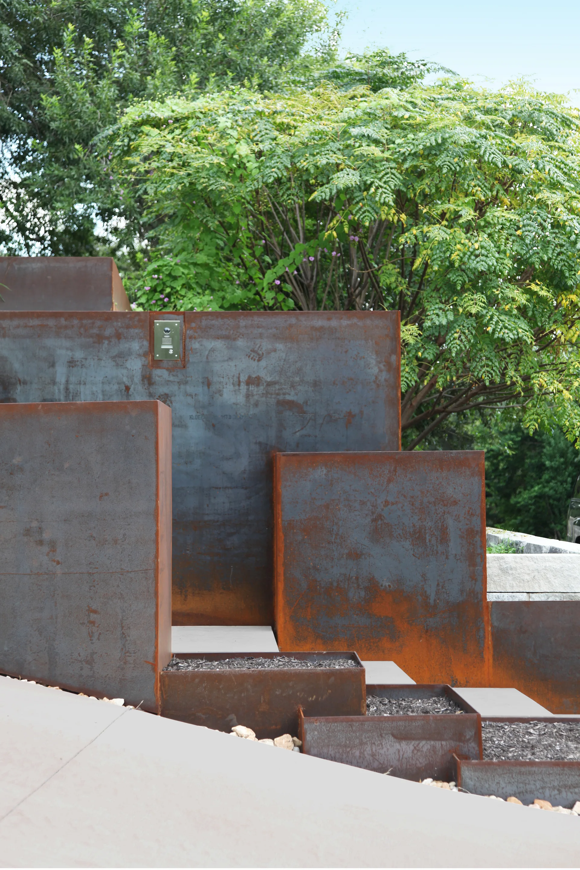 A stepped outdoor landscape features weathered Corten steel retaining walls and rectangular planter boxes set against lush green trees.