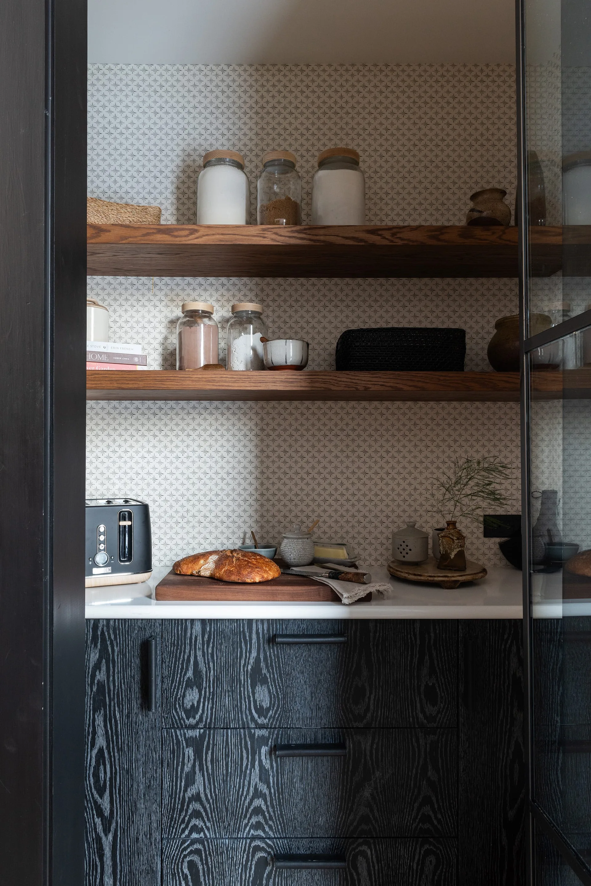 A kitchen pantry features dark wood grain cabinets, a white countertop, patterned wallpaper, and floating wooden shelves with glass storage jars.
