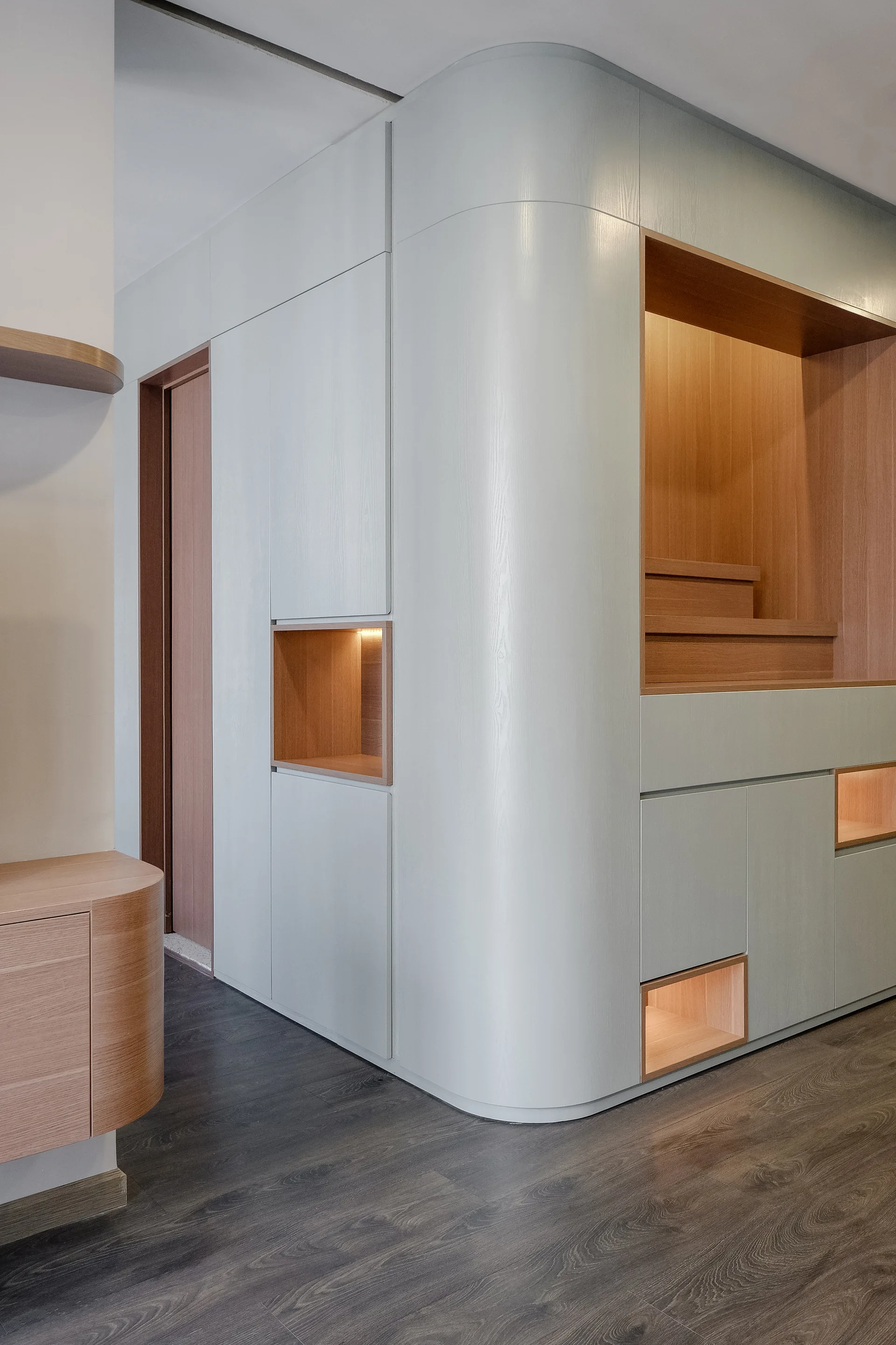 Custom pale grey cabinetry with a rounded corner features integrated wood-lined niches illuminated with warm light.