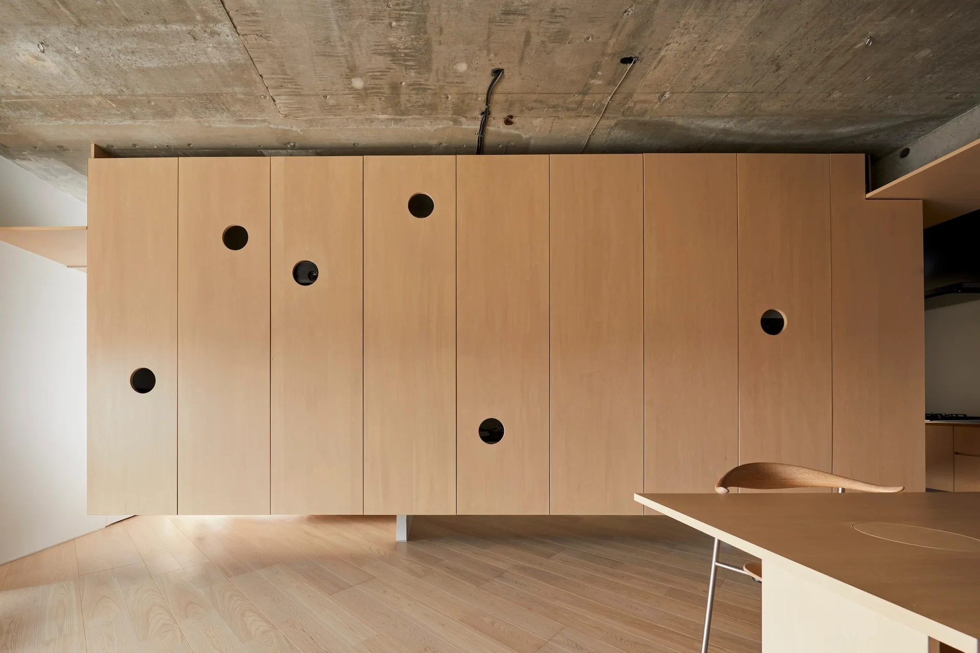 A minimalist dining area featuring a large light wood cabinet wall with circular cutouts under an exposed raw concrete ceiling.