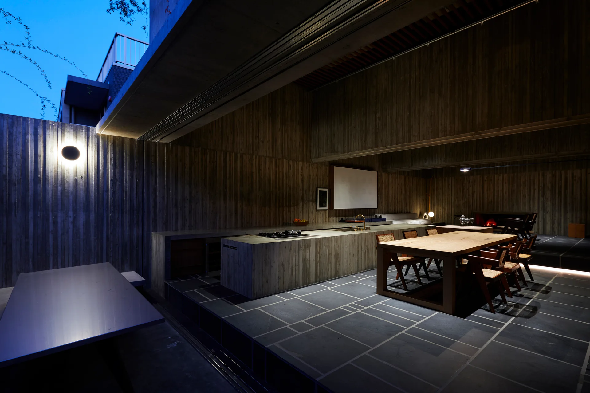 A modern brutalist kitchen features board-formed concrete walls, a dark grid-patterned stone floor, a concrete island, and a wooden dining table.