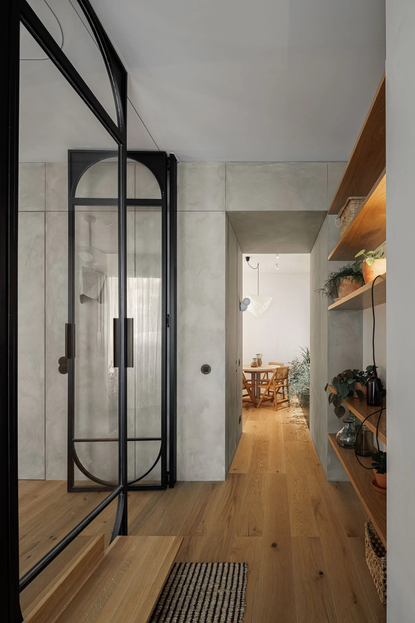 A minimalist hallway features concrete-look walls, a black-framed arched glass door, and wooden floating shelves displaying potted plants.
