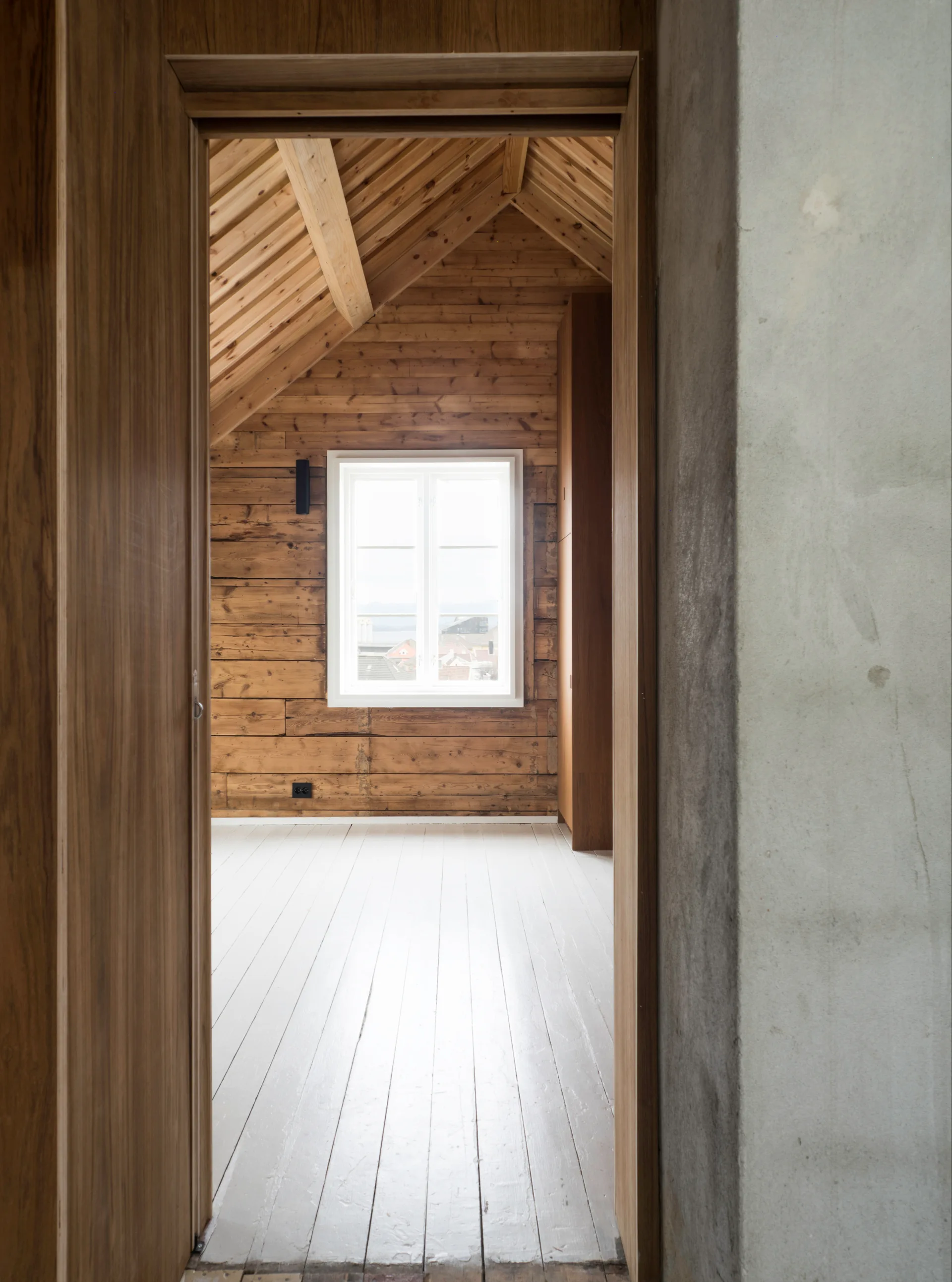 A view through a wooden doorway into an empty attic room with horizontal wood paneled walls, a sloped ceiling, and painted white floorboards.