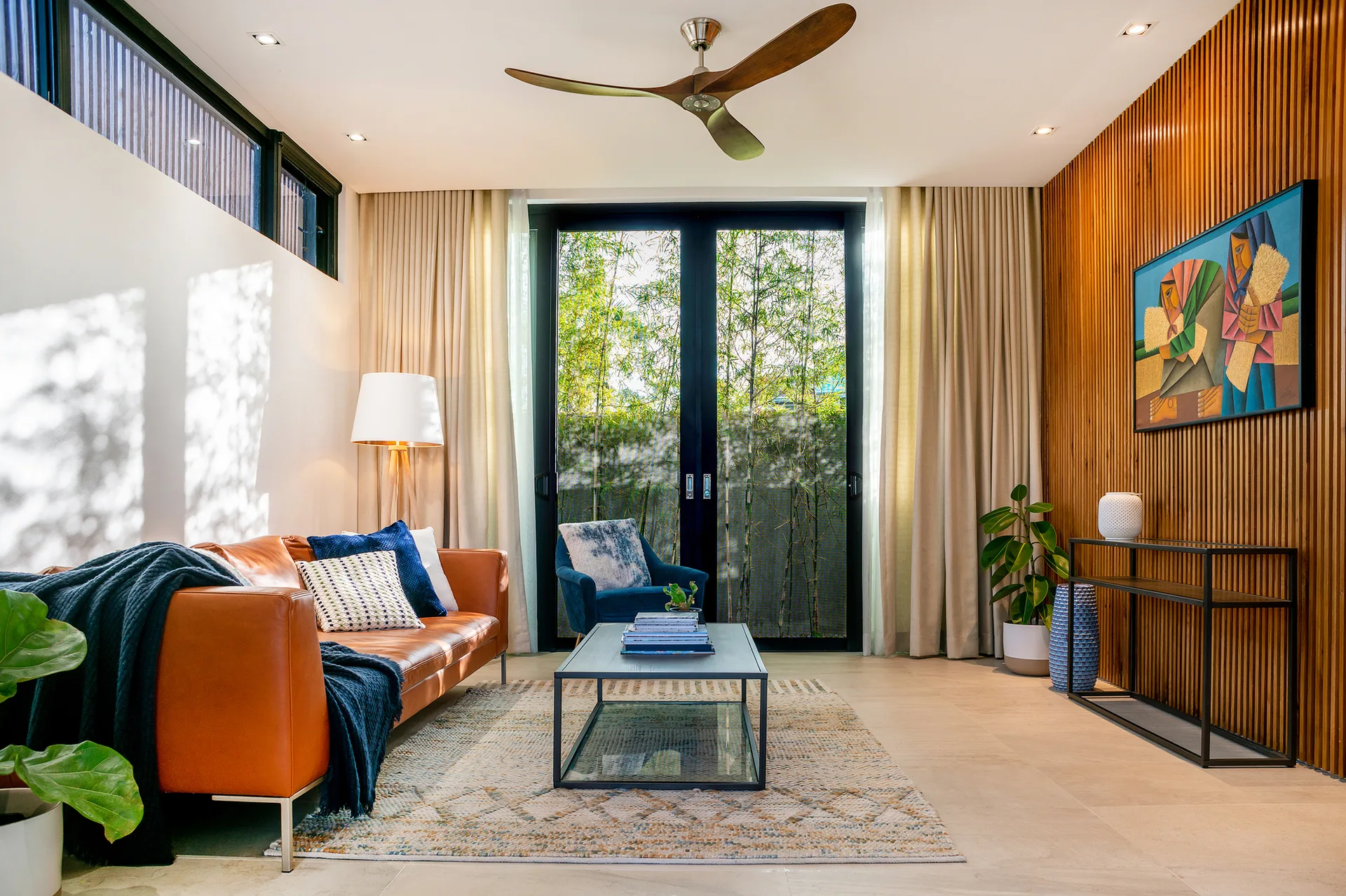 A modern living room features a tan leather sofa, a vertical wood slat accent wall, and sliding glass doors opening to outdoor greenery.