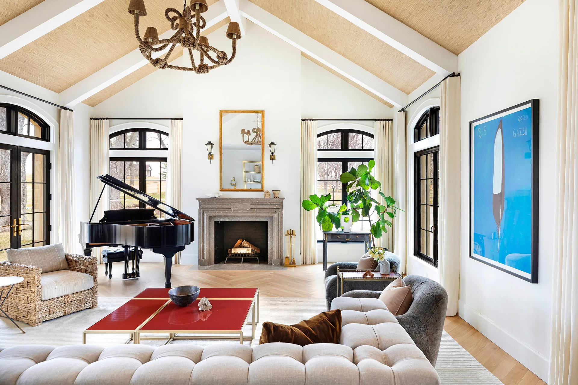A spacious white living room features a vaulted ceiling with tan textured panels, a central stone fireplace, a black grand piano, and a tufted sectional.