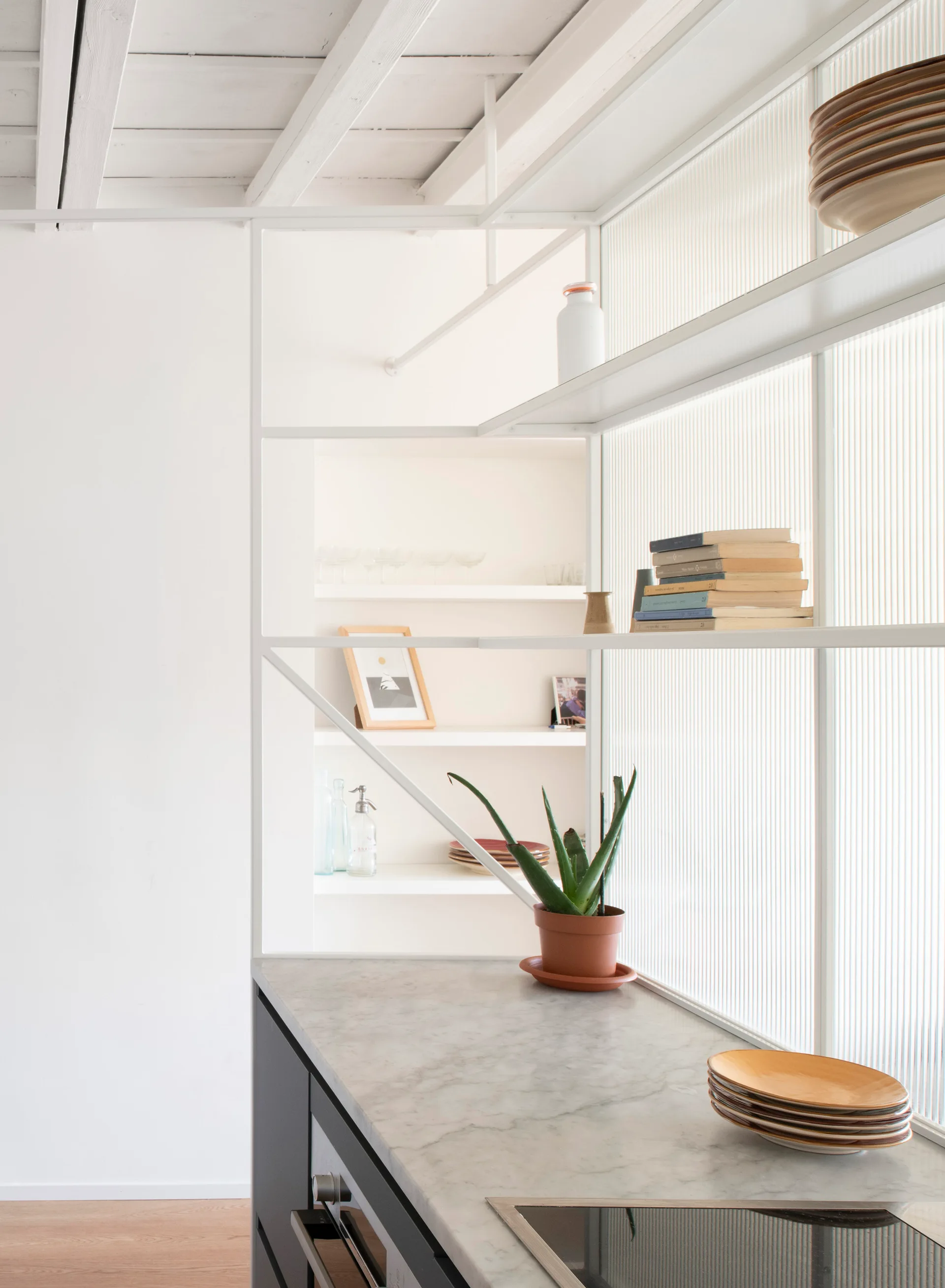 A modern kitchen features a custom white metal shelving unit with reeded glass panels above a marble-topped counter.
