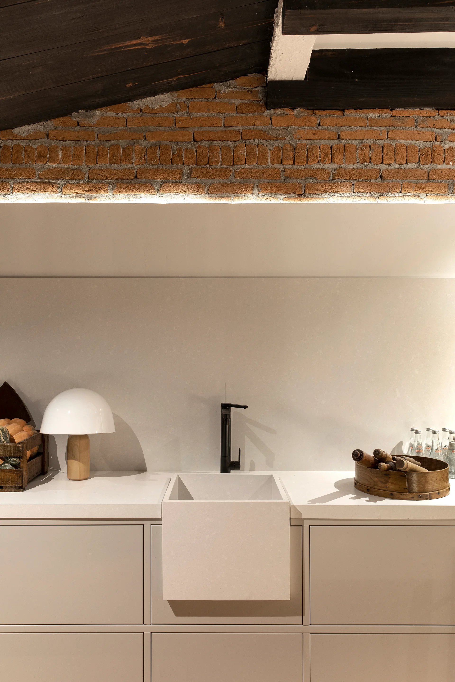 A beige kitchen counter features a blocky stone farmhouse sink, a black faucet, flat-front cabinetry, and an exposed brick wall under dark ceiling beams.