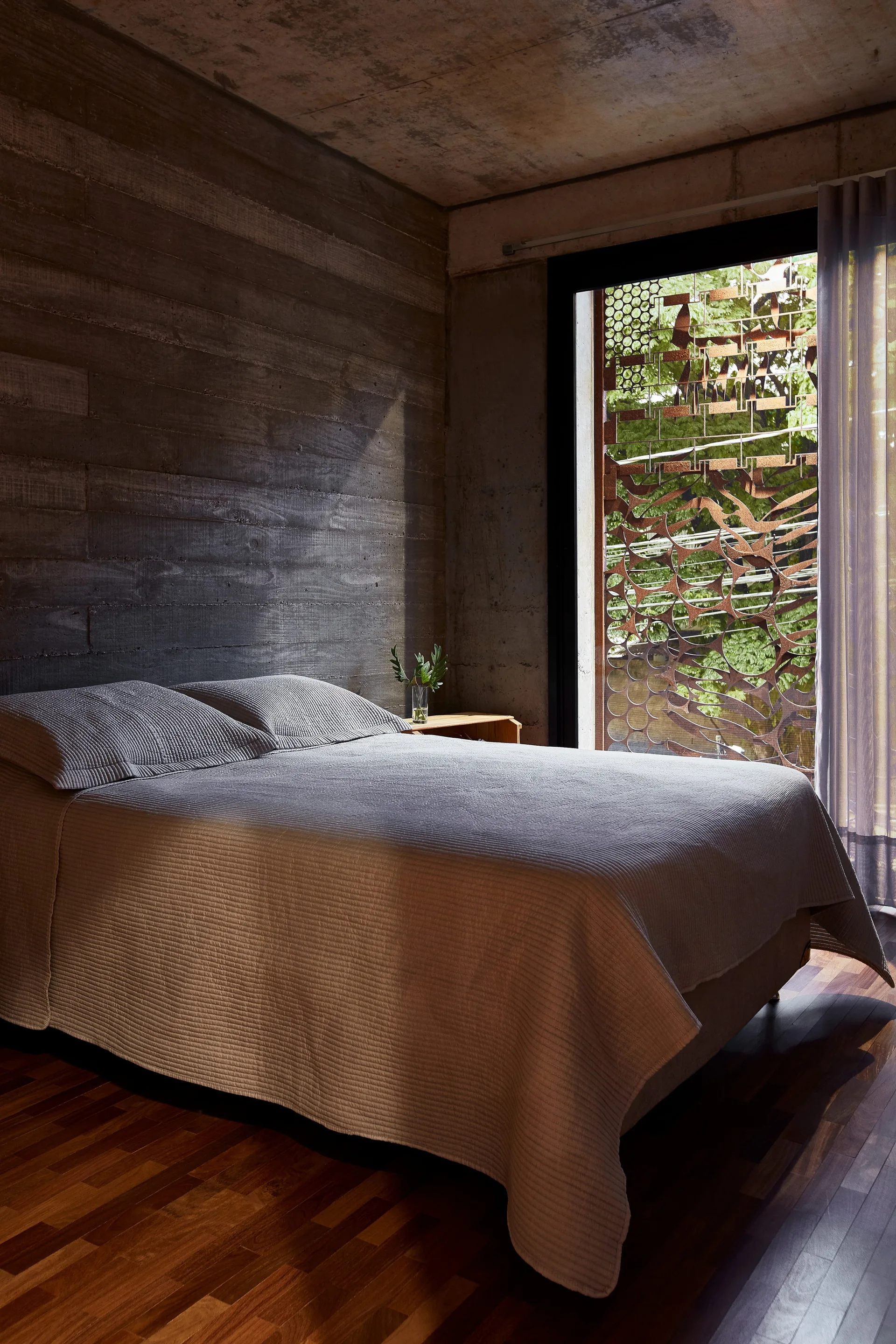 A minimalist bedroom featuring a bed with grey bedding set against a board-formed concrete wall next to a window with a patterned metal screen.