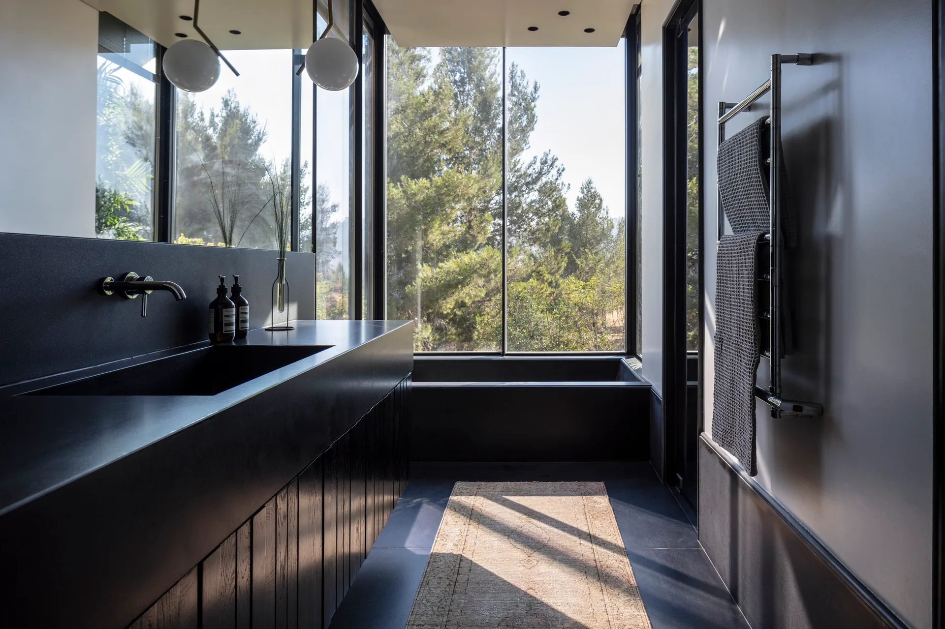 A modern black bathroom with a dark slatted vanity, a built-in tub under large panoramic windows overlooking trees, and a woven floor runner.