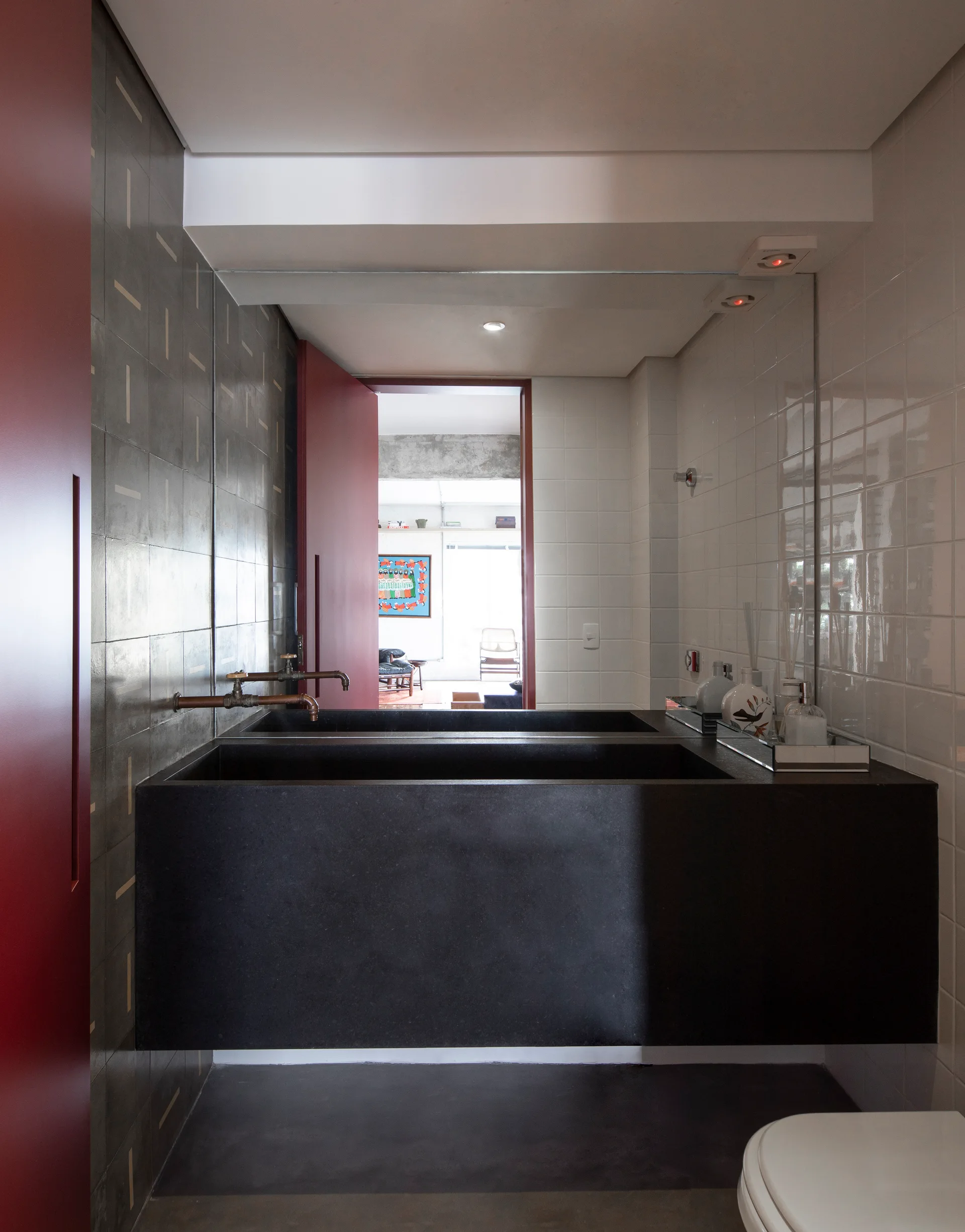 A modern bathroom features a large floating black stone sink with industrial copper faucets, set between a grey tiled wall and a glossy white tiled wall.