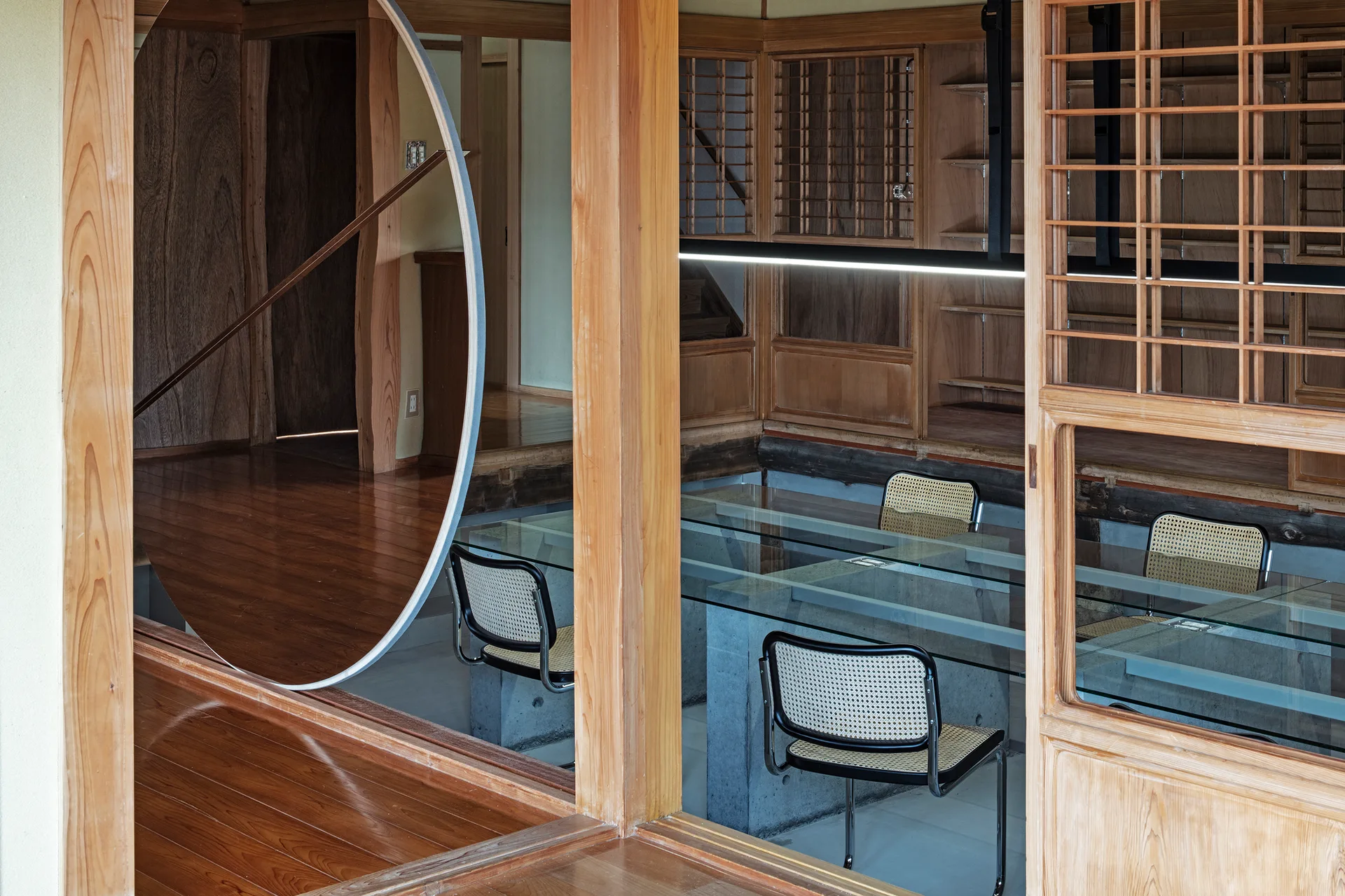 A modern workspace features a glass-top table on concrete bases, cane-backed chairs, and traditional wood lattice panels with a large circular mirror.
