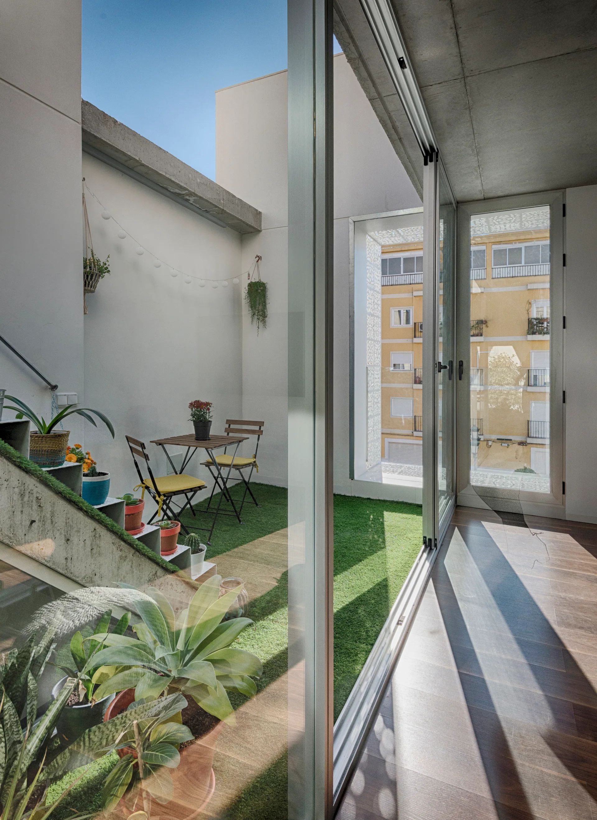 A small urban terrace featuring green artificial turf, a folding bistro set with yellow cushions, and potted plants on concrete stairs.