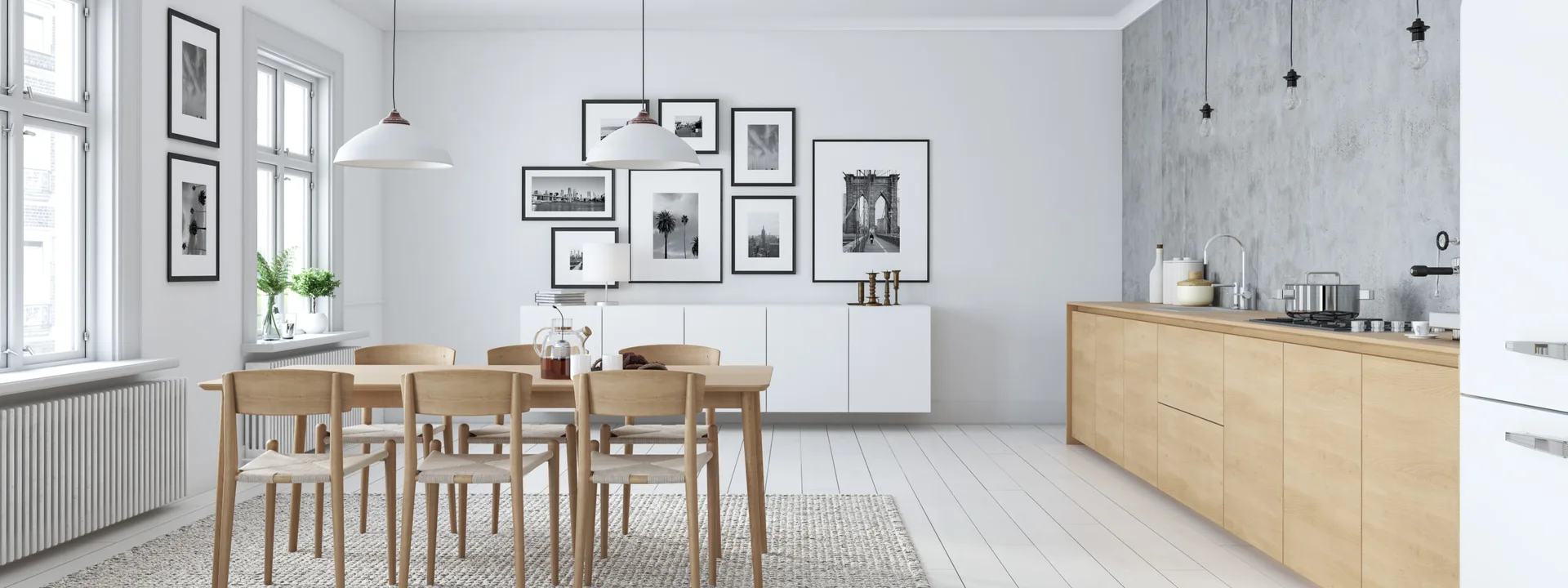 A bright dining room and kitchen featuring a light wood table, a gallery wall above a white sideboard, and a grey concrete-effect accent wall.