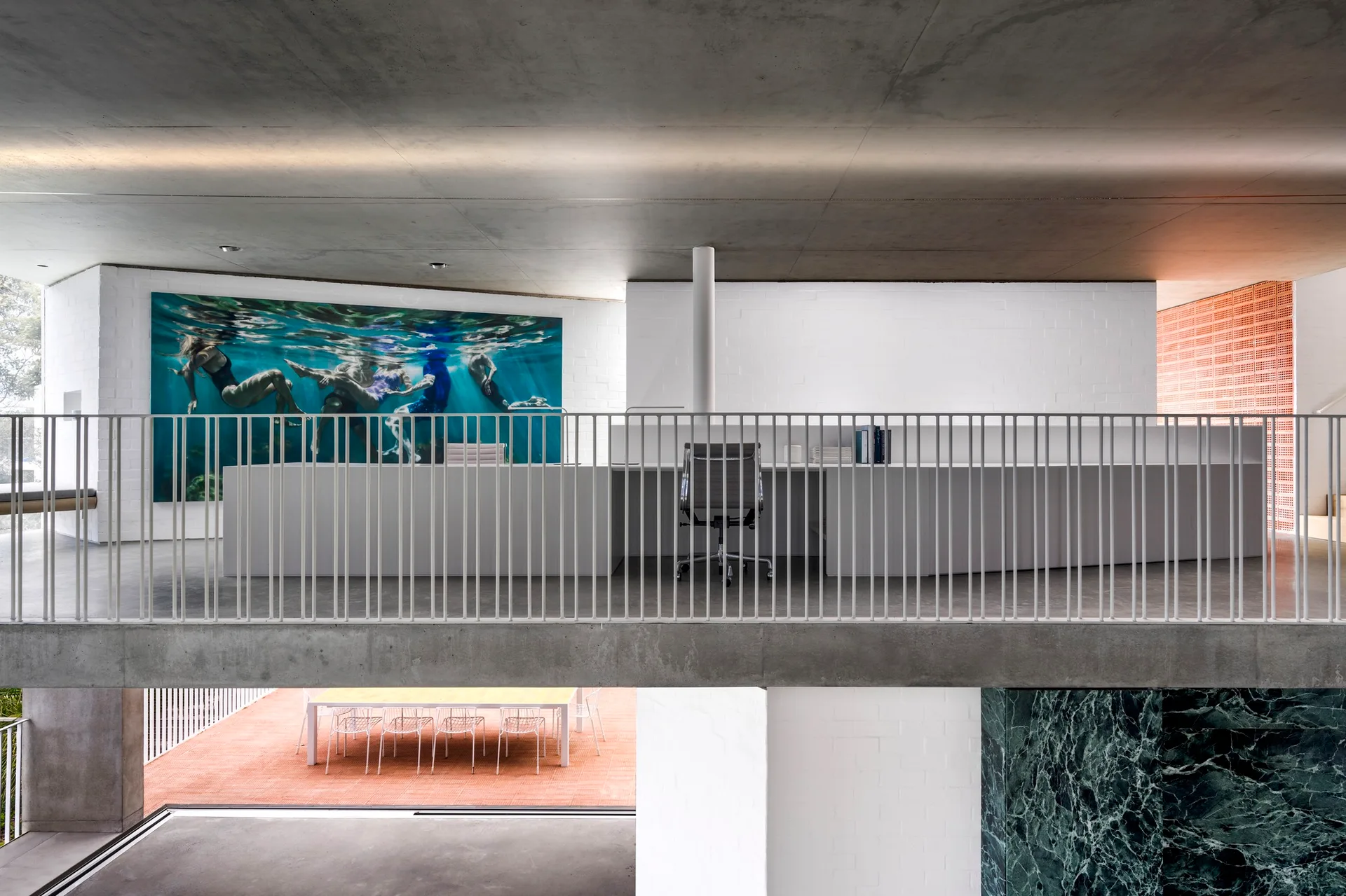 A minimalist mezzanine workspace features a long white desk, a grey office chair, a vertical metal railing, and a large underwater art print.