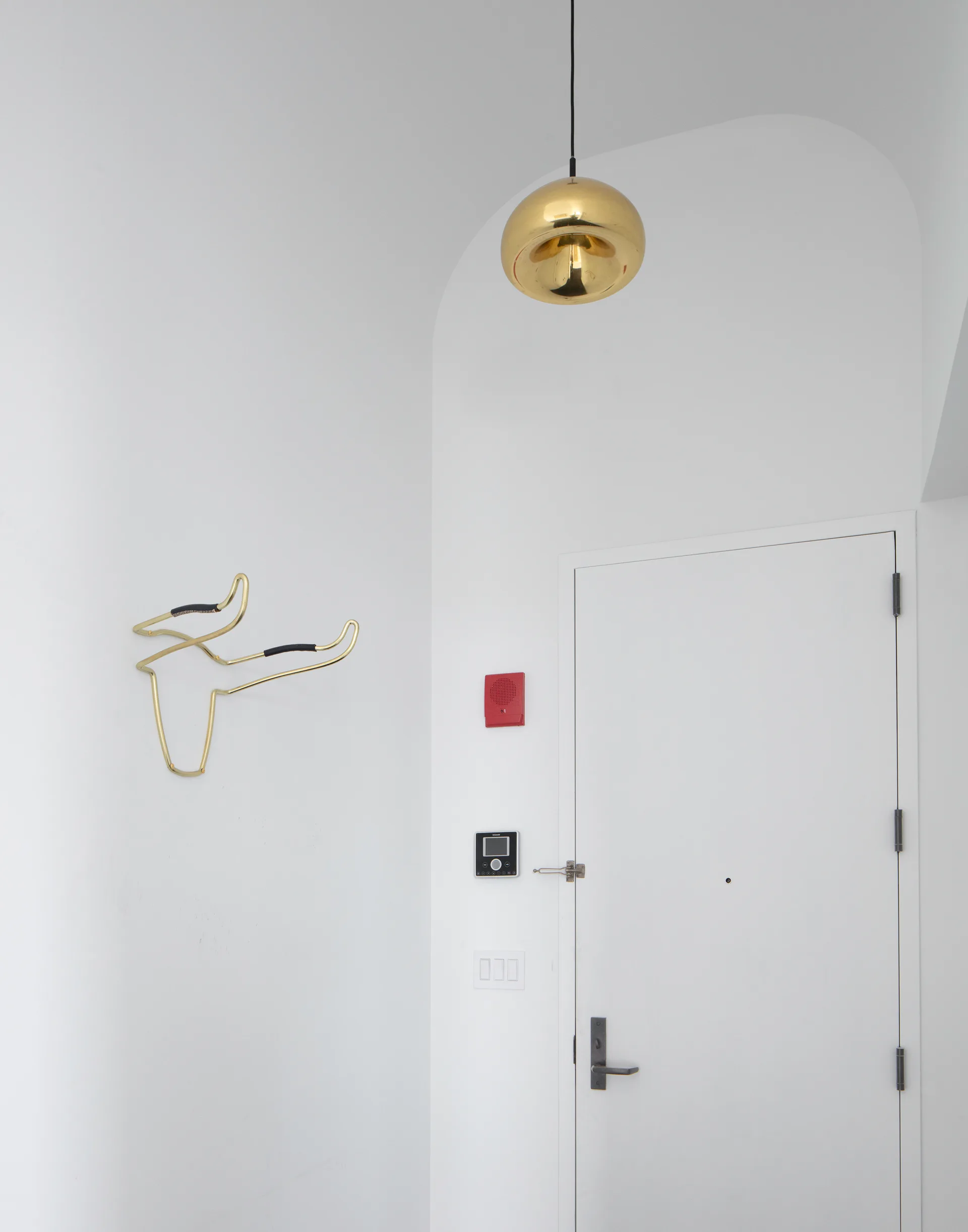 A minimalist white entryway features a shiny gold dome pendant light, an arched wall, and a sculptural gold wire hook on a smooth plaster wall.