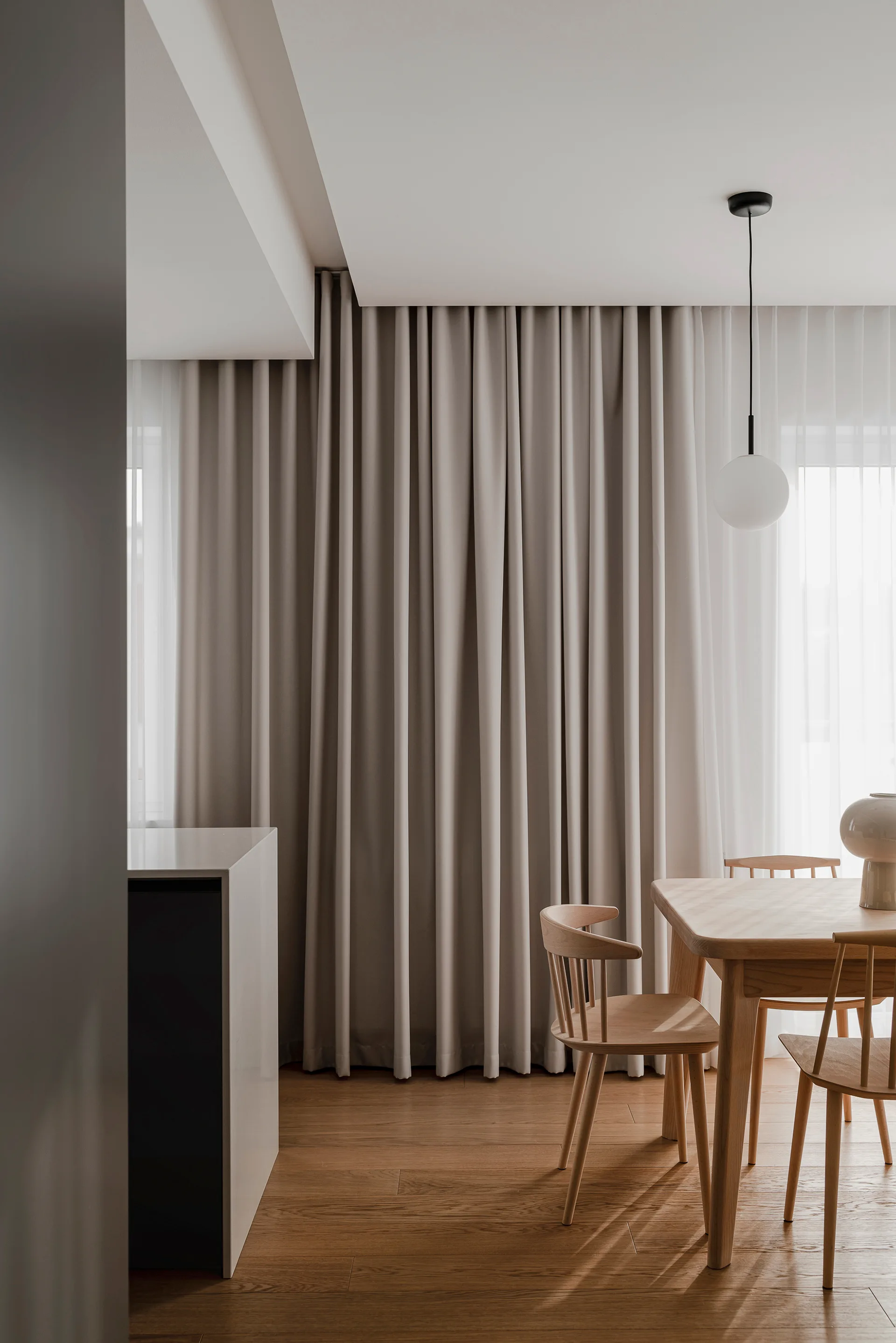 A minimalist dining room features a light wood table and spindle-back chairs set against full-length beige curtains and a white pendant light.