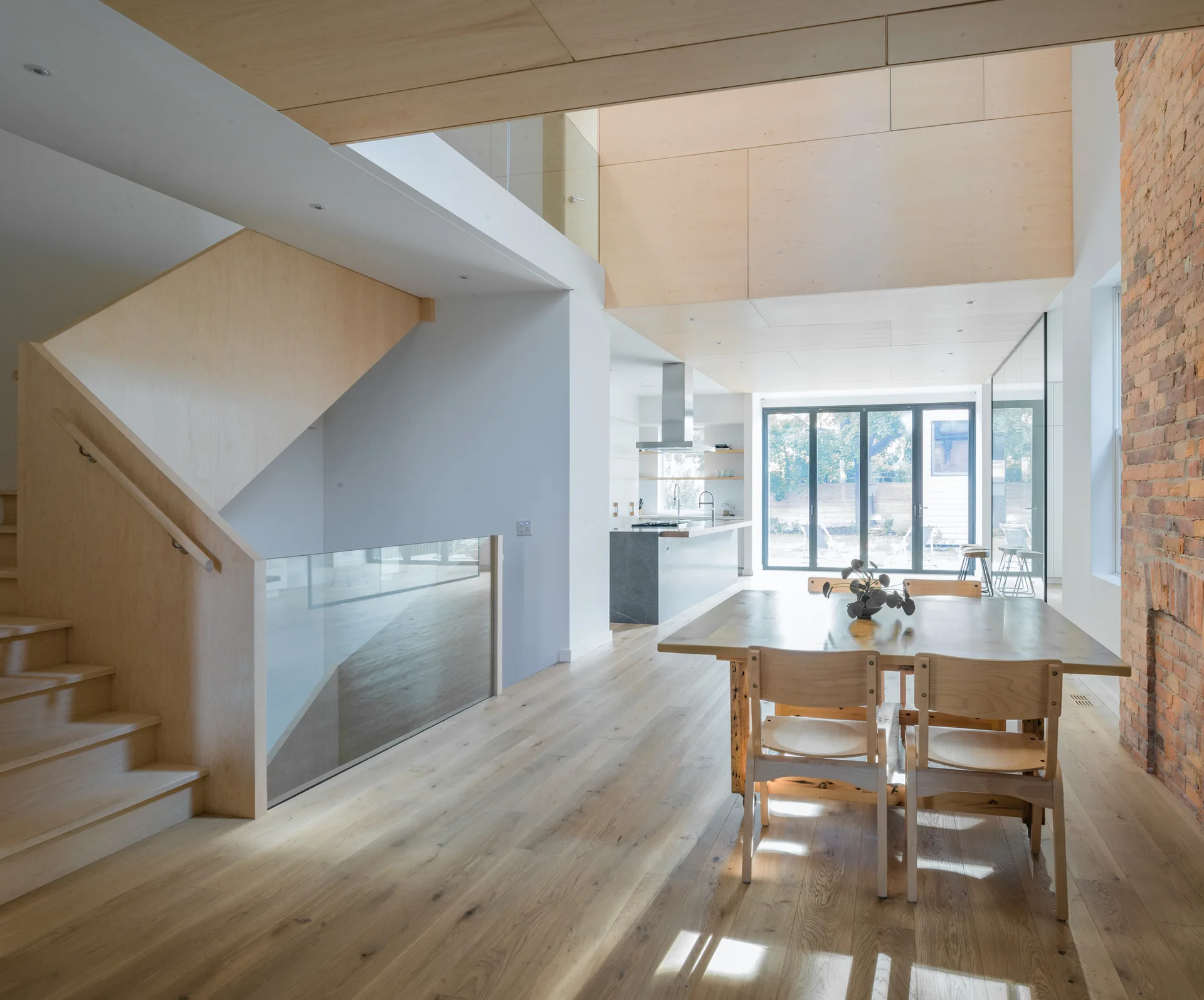 A dining area with a wooden table, a light plywood staircase with a glass railing, and a tall exposed brick chimney.