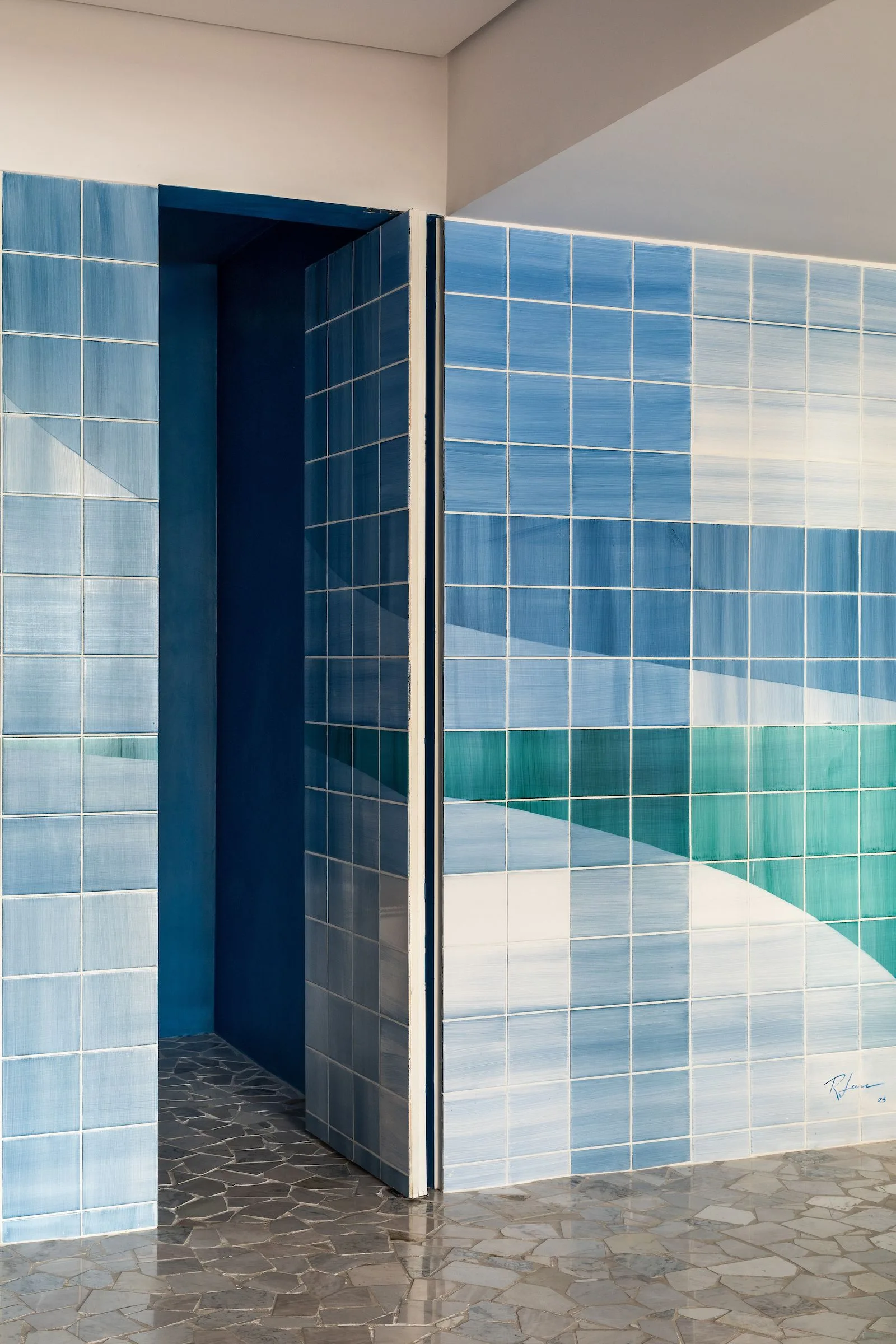 A corner of a room featuring a blue and white geometric tile wall mural, an open doorway, and a grey stone mosaic floor.