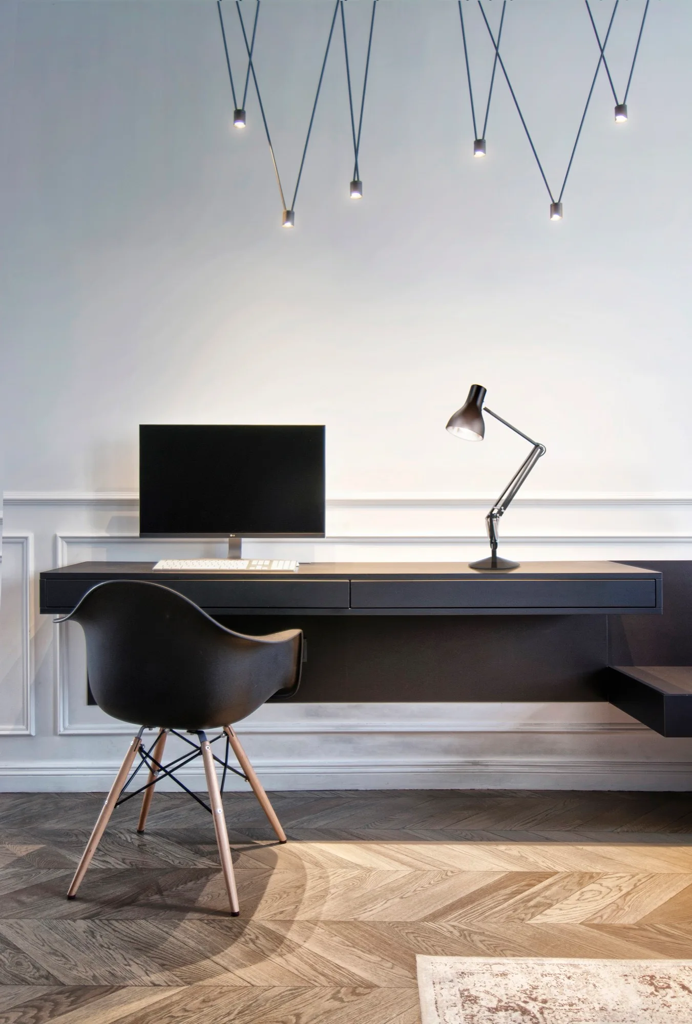 A minimalist black floating desk is mounted on a white wainscoted wall, paired with a molded black chair on herringbone wood flooring.