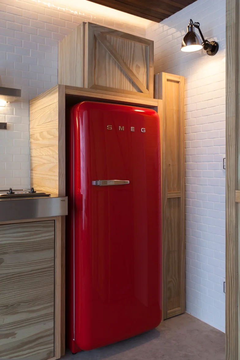 A bright red retro refrigerator is built into a custom light wood frame against a white subway tile wall with a black task light overhead.