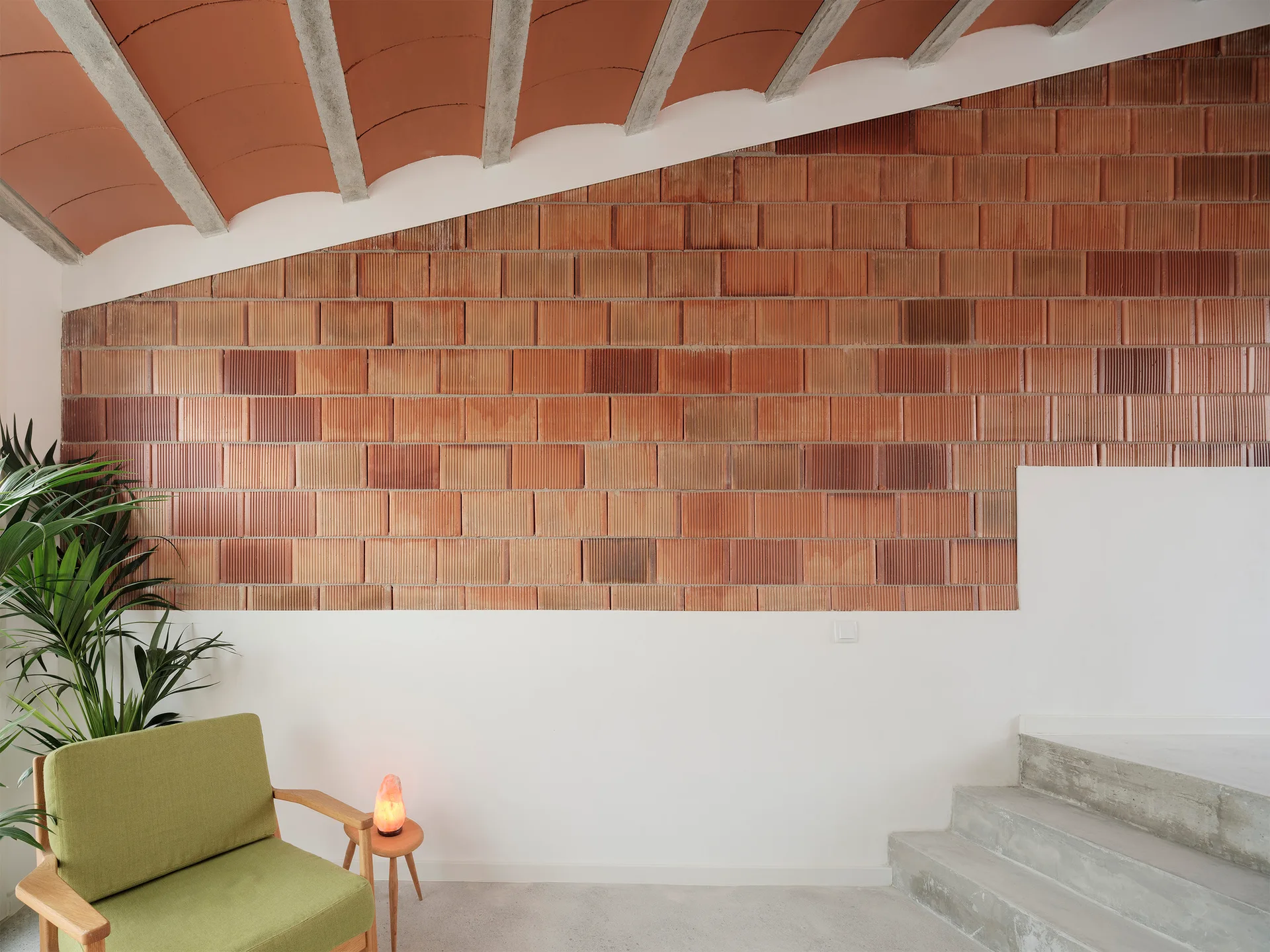 A minimalist staircase landing features a vaulted terracotta ceiling, a ribbed brick wall, concrete stairs, and a green armchair with a small side table.