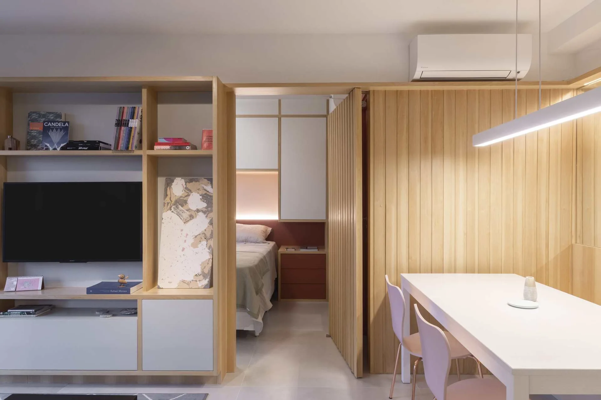 A minimalist studio apartment features custom light wood built-in cabinetry, a white dining table with chairs, and a slatted wood sliding door.
