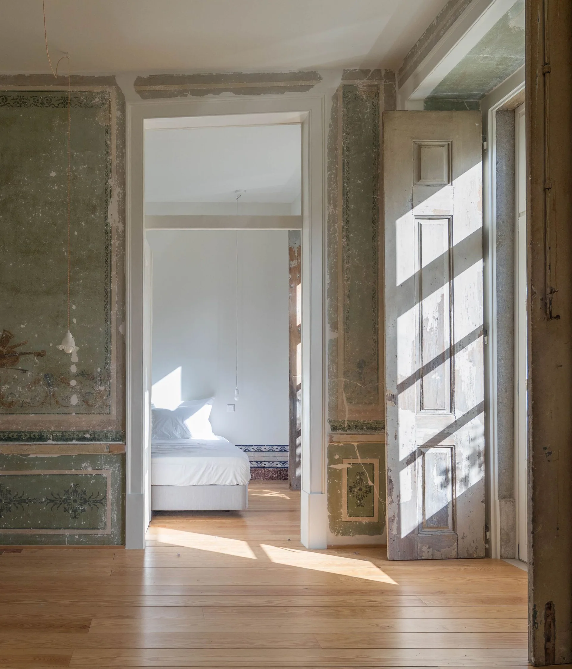 A view through an open doorway of a minimalist white bedroom, contrasted by distressed sage green plaster walls and light wood flooring in the foreground.