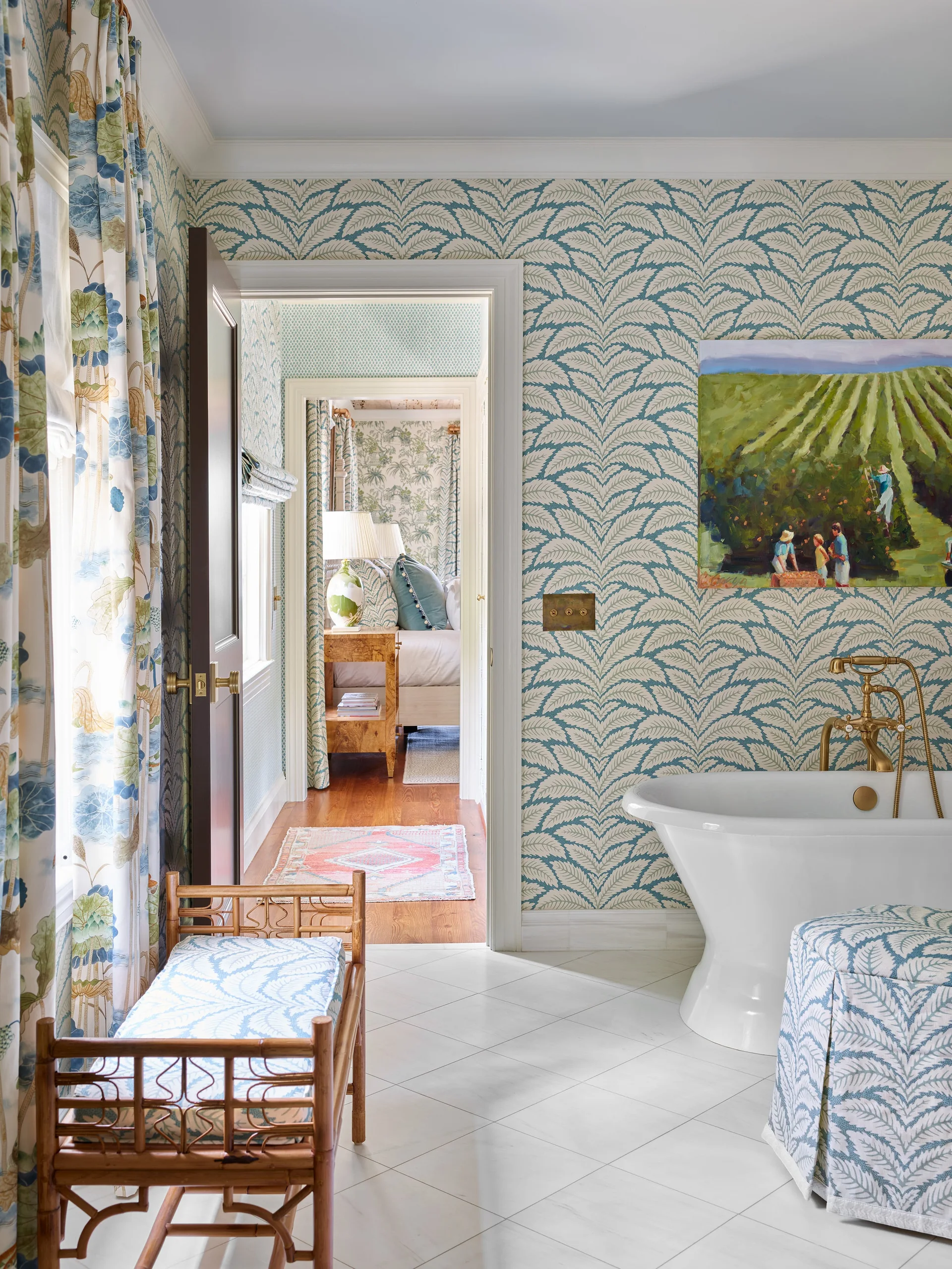 A bathroom with blue leaf-patterned wallpaper, a white freestanding tub, a bamboo bench, and an open doorway to a bedroom.