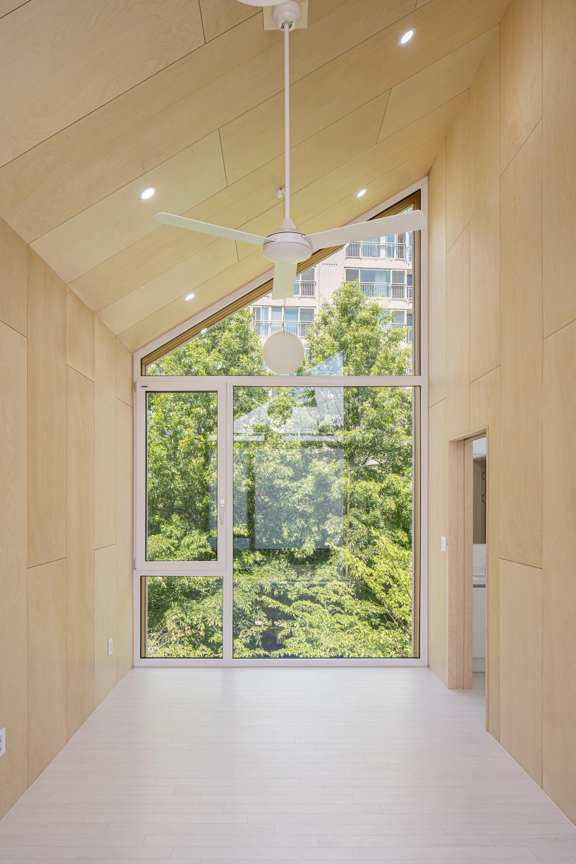 A minimalist room features light plywood paneled walls and a slanted ceiling, centered around a large window showing green trees outside.