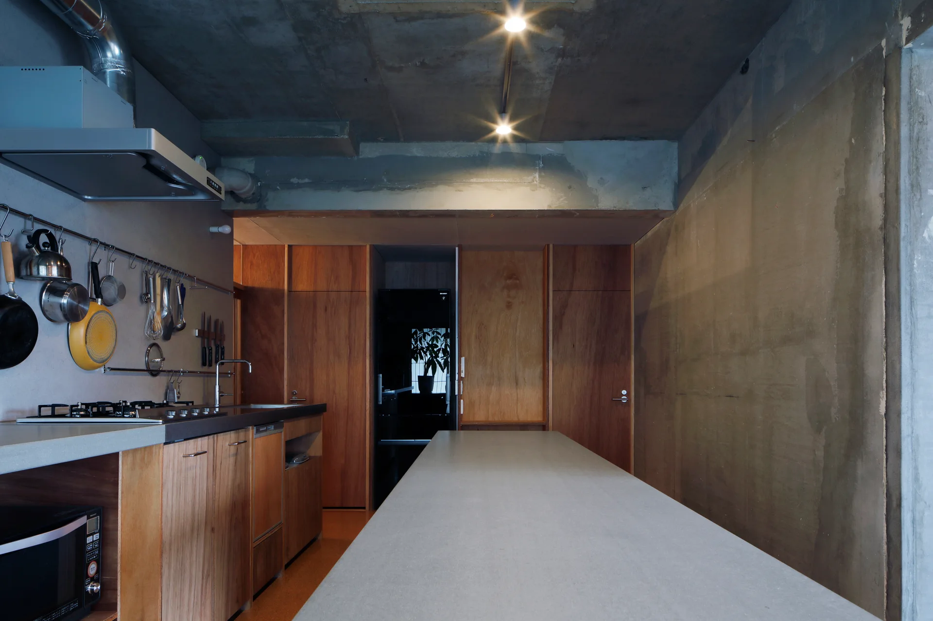 An industrial kitchen features a raw concrete ceiling, warm plywood cabinetry, a long grey island, and a stainless steel wall organizer for cookware.