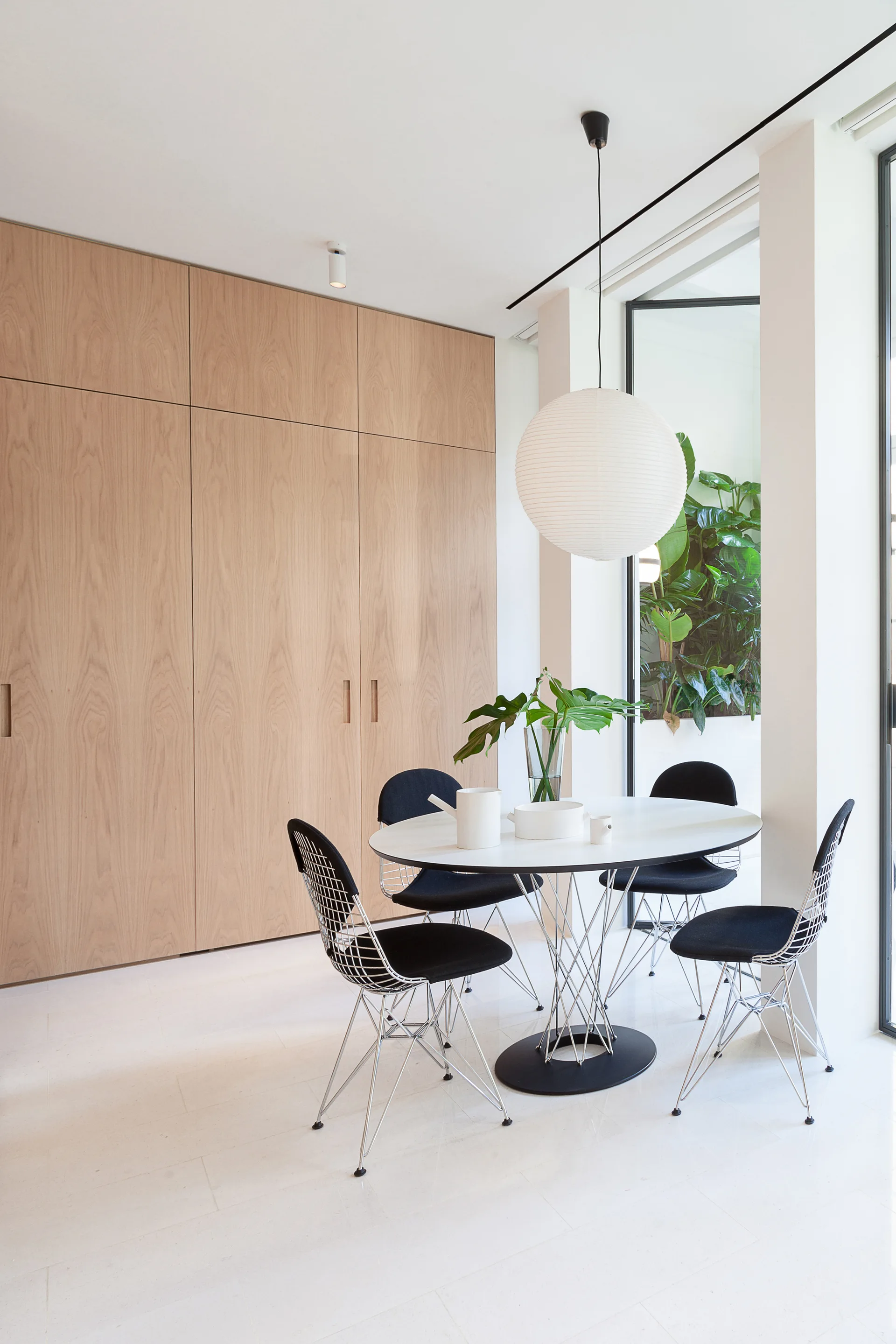 A minimalist dining area featuring a white oval table, black wire chairs, a large paper pendant light, and a floor-to-ceiling wood cabinet wall.