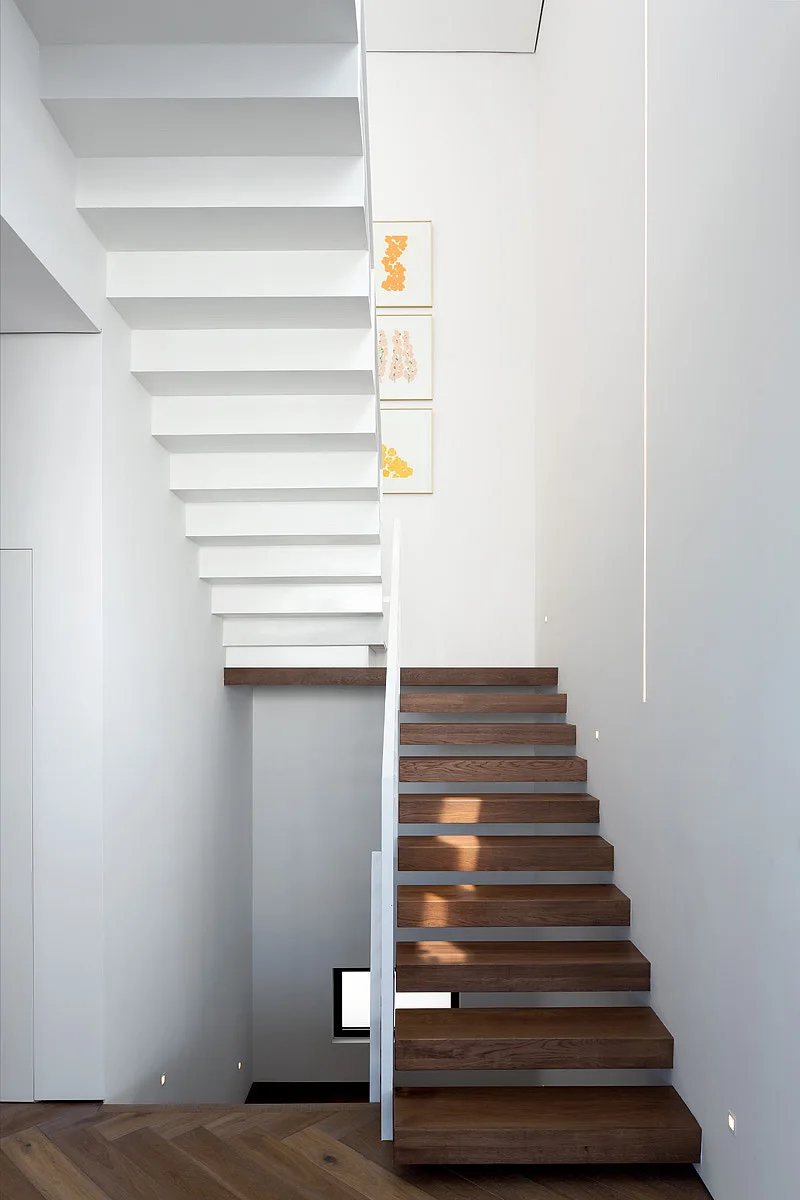 A minimalist stairwell features floating dark wood treads, white metal railings, herringbone wood floors, and small integrated wall lights.