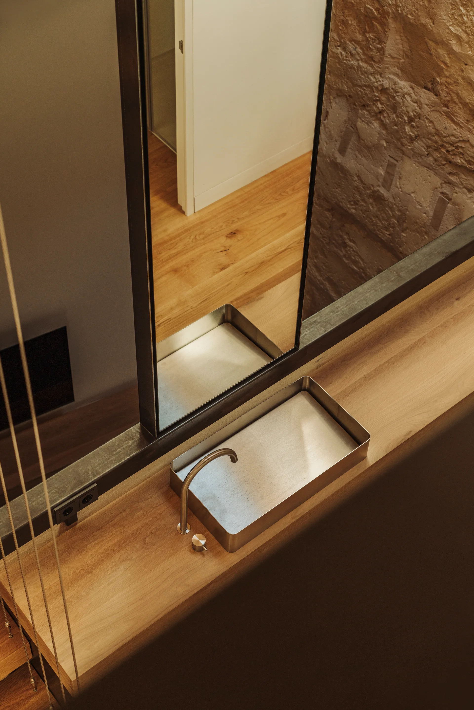 A high-angle view of a minimalist metal sink on a wood countertop in a bathroom next to a large mirror and an exposed stone wall.