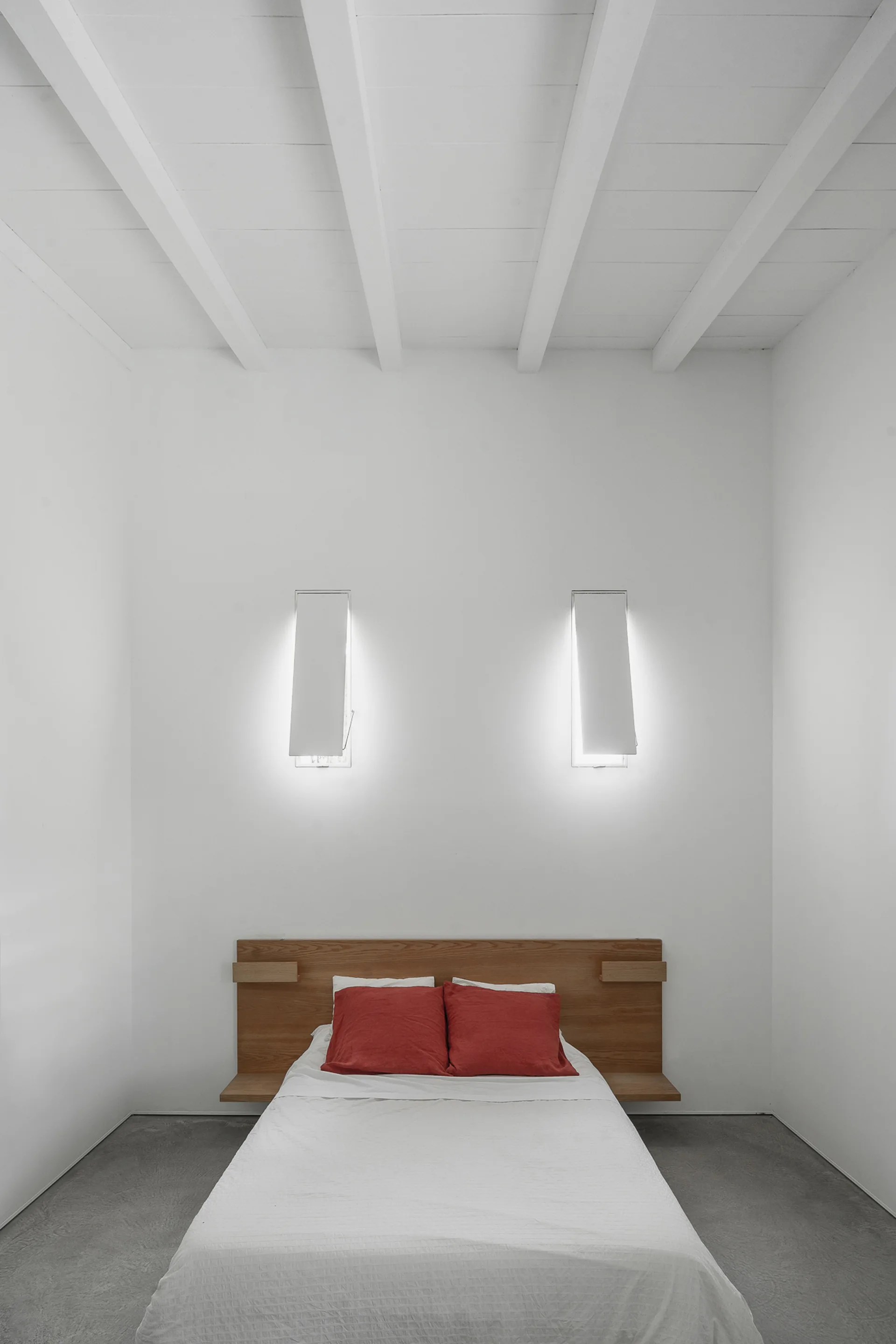 A minimalist bedroom features a low bed with an integrated wooden headboard, red pillows, and glowing wall sconces under white ceiling beams.