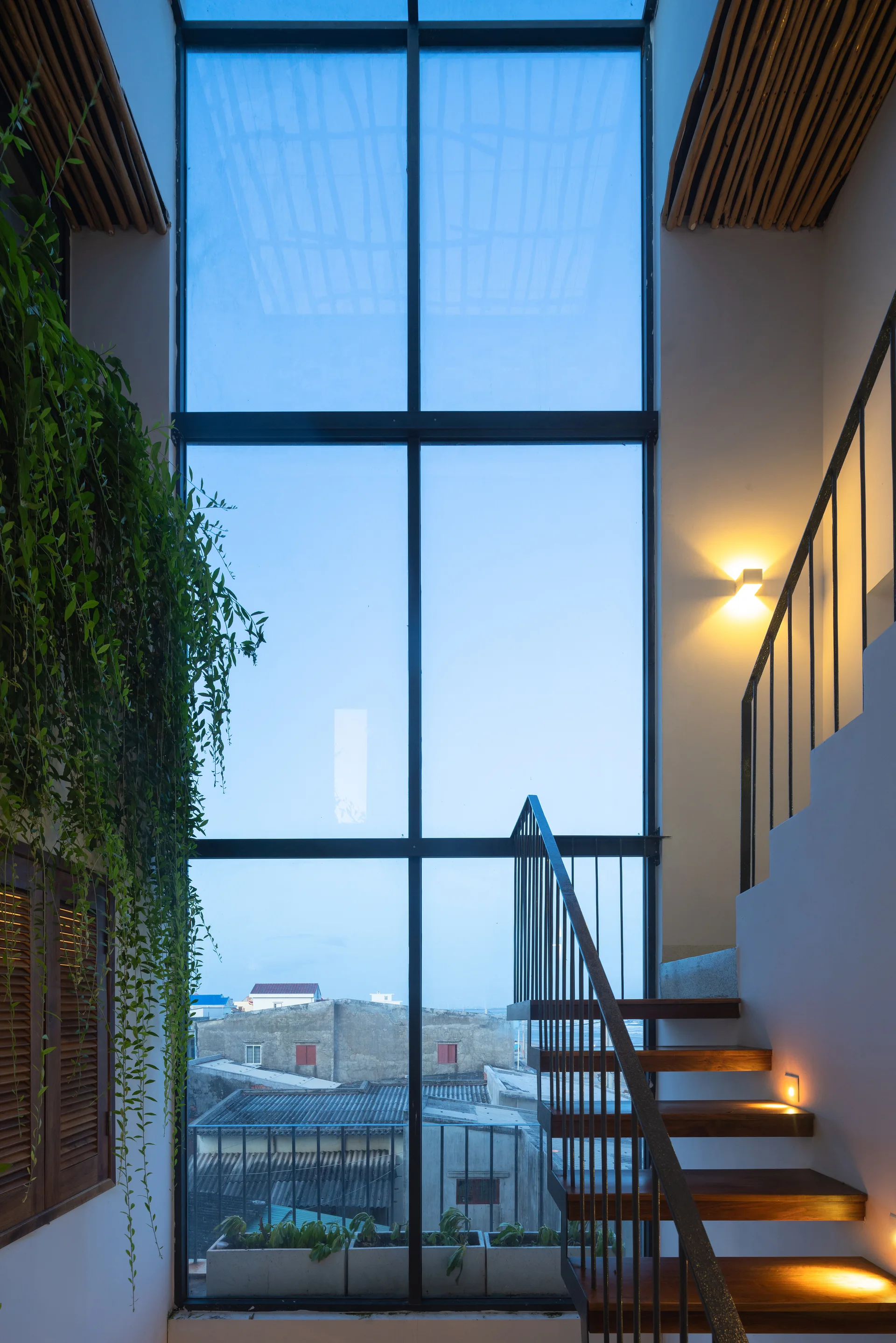 A wooden staircase with integrated tread lighting stands next to a tall grid window and a wall covered in trailing green plants.
