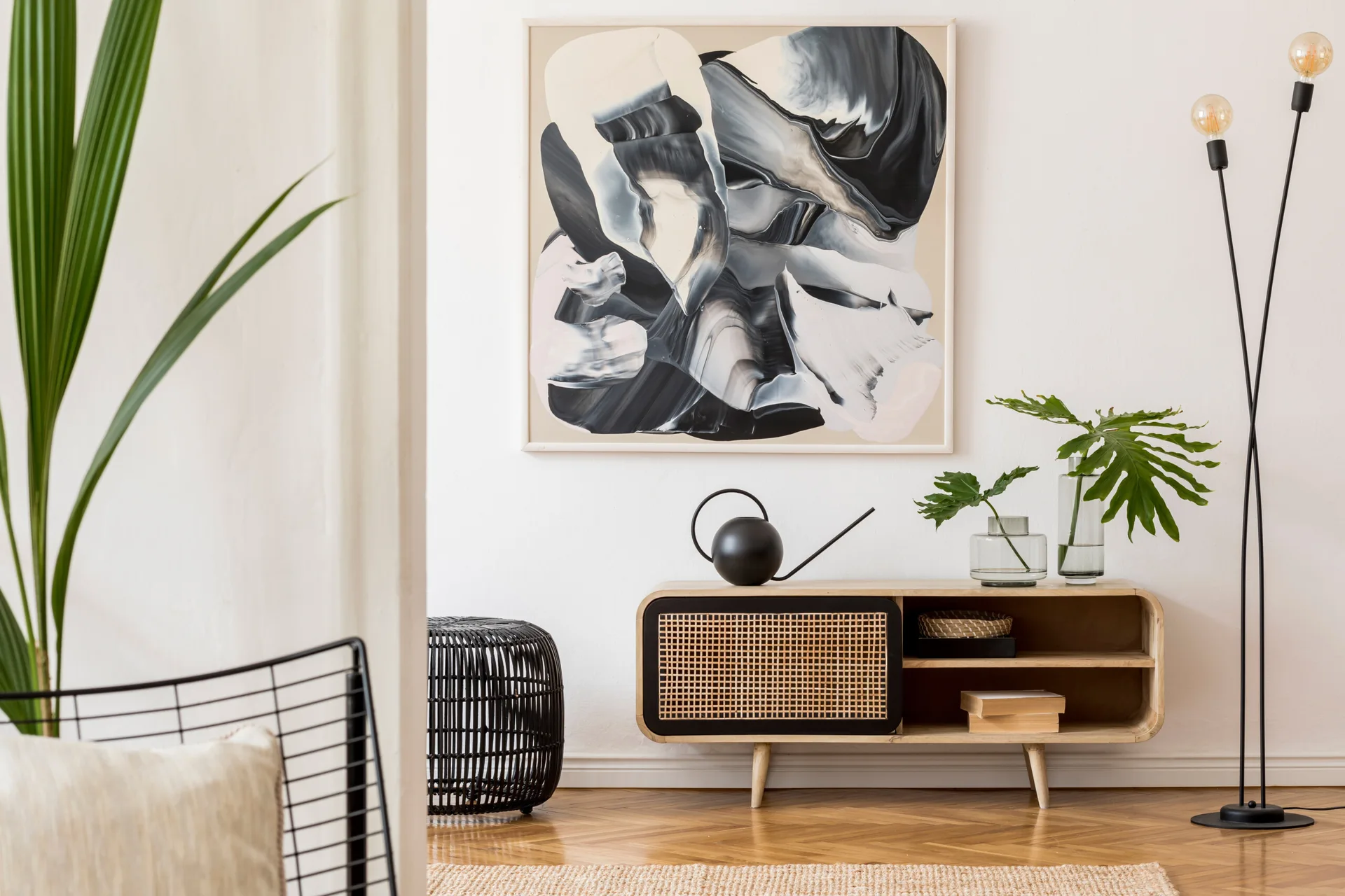 A wooden sideboard with a woven rattan door sits beneath a large black and white abstract painting in a minimalist room with herringbone floors.