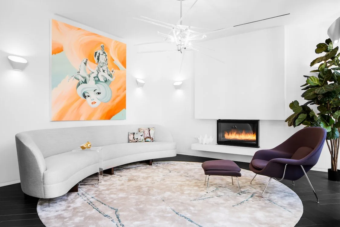 A contemporary white living room features a curved light-colored sofa, a deep purple armchair with an ottoman, a modern fireplace, and large orange artwork.