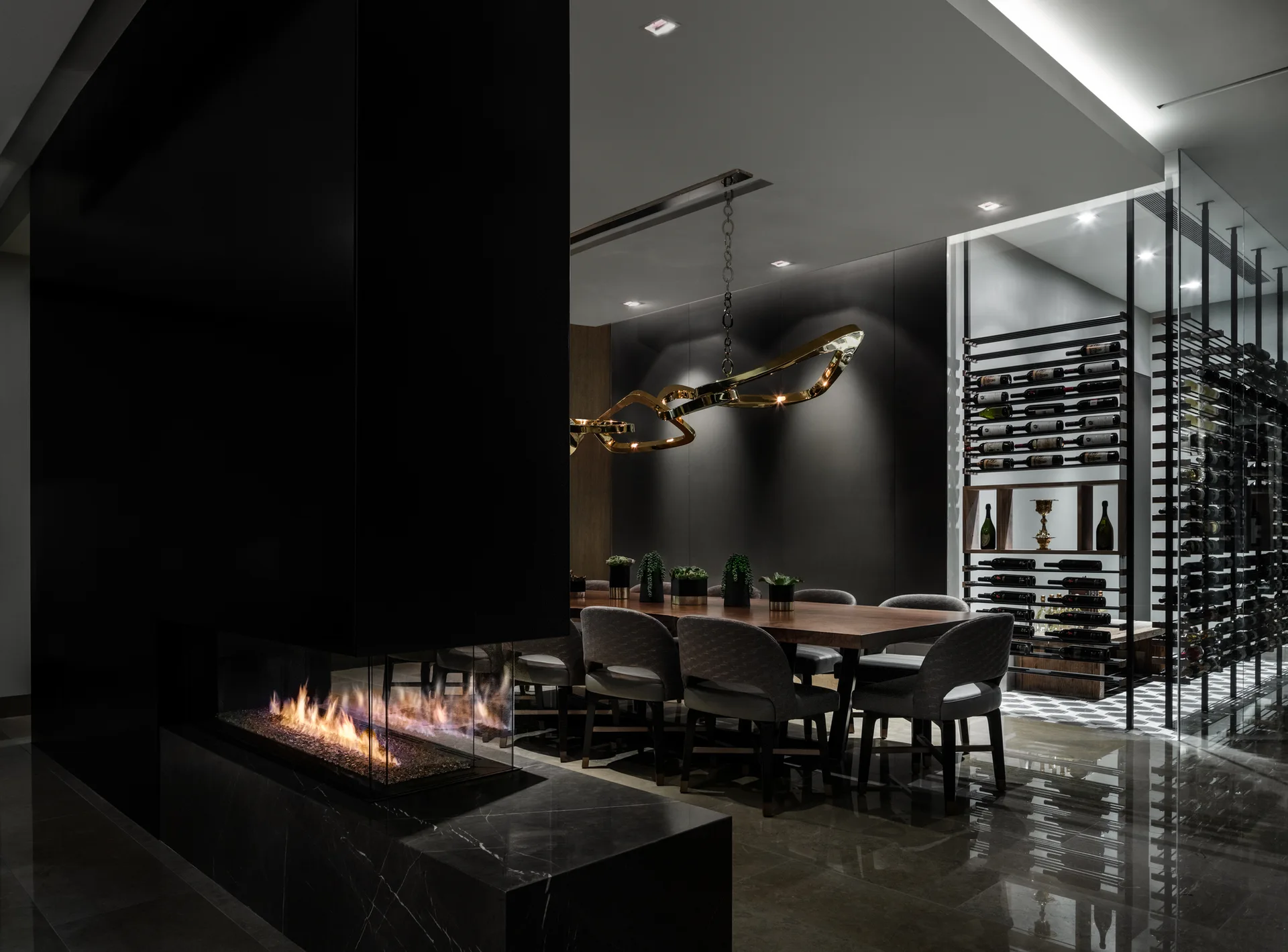 A contemporary dining room features a long wooden table, grey chairs, a sculptural gold chandelier, a linear fireplace, and an adjacent glass wine display.