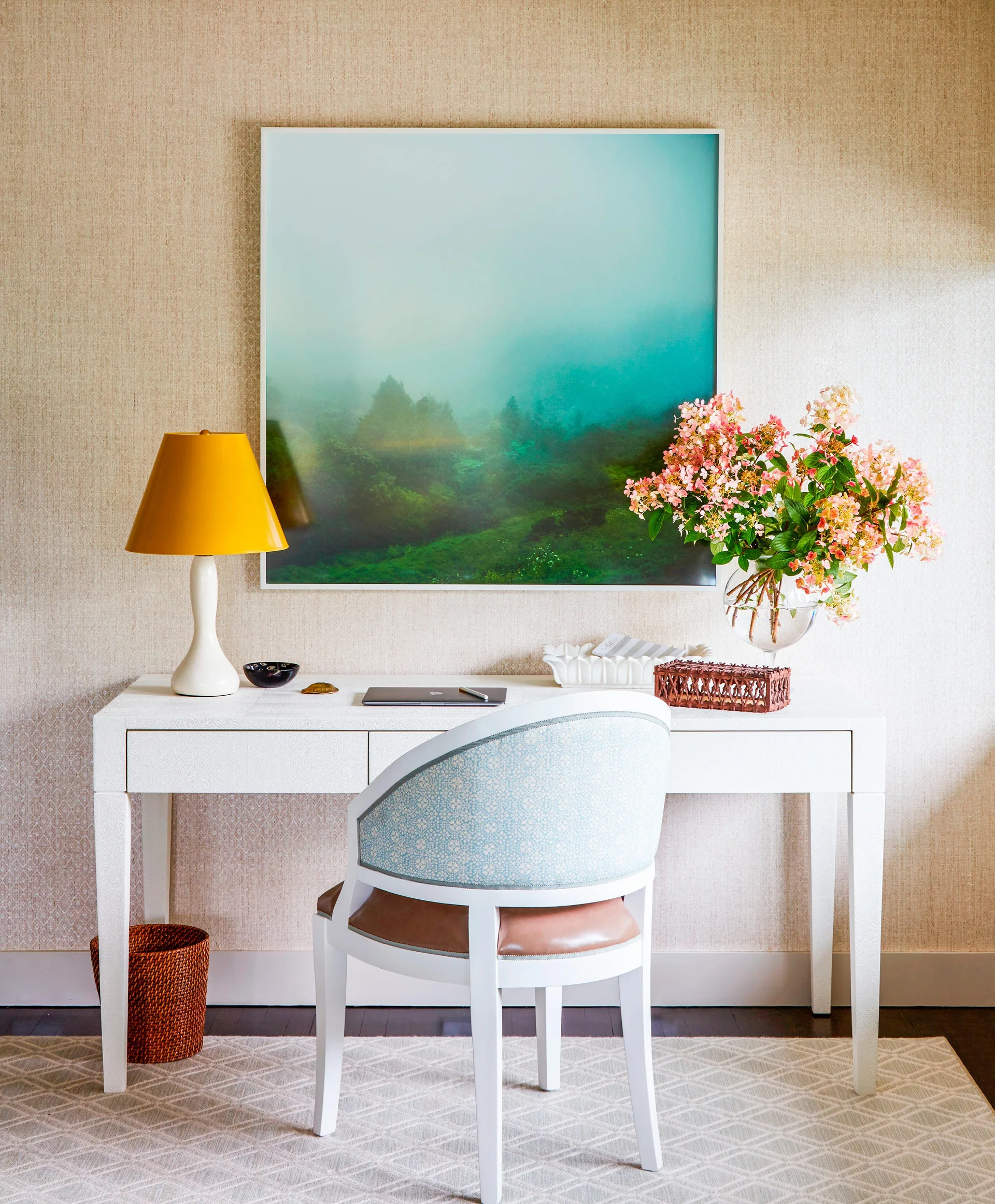 A white writing desk with a yellow table lamp, glass vase of pink flowers, and patterned armchair against textured wallpaper.