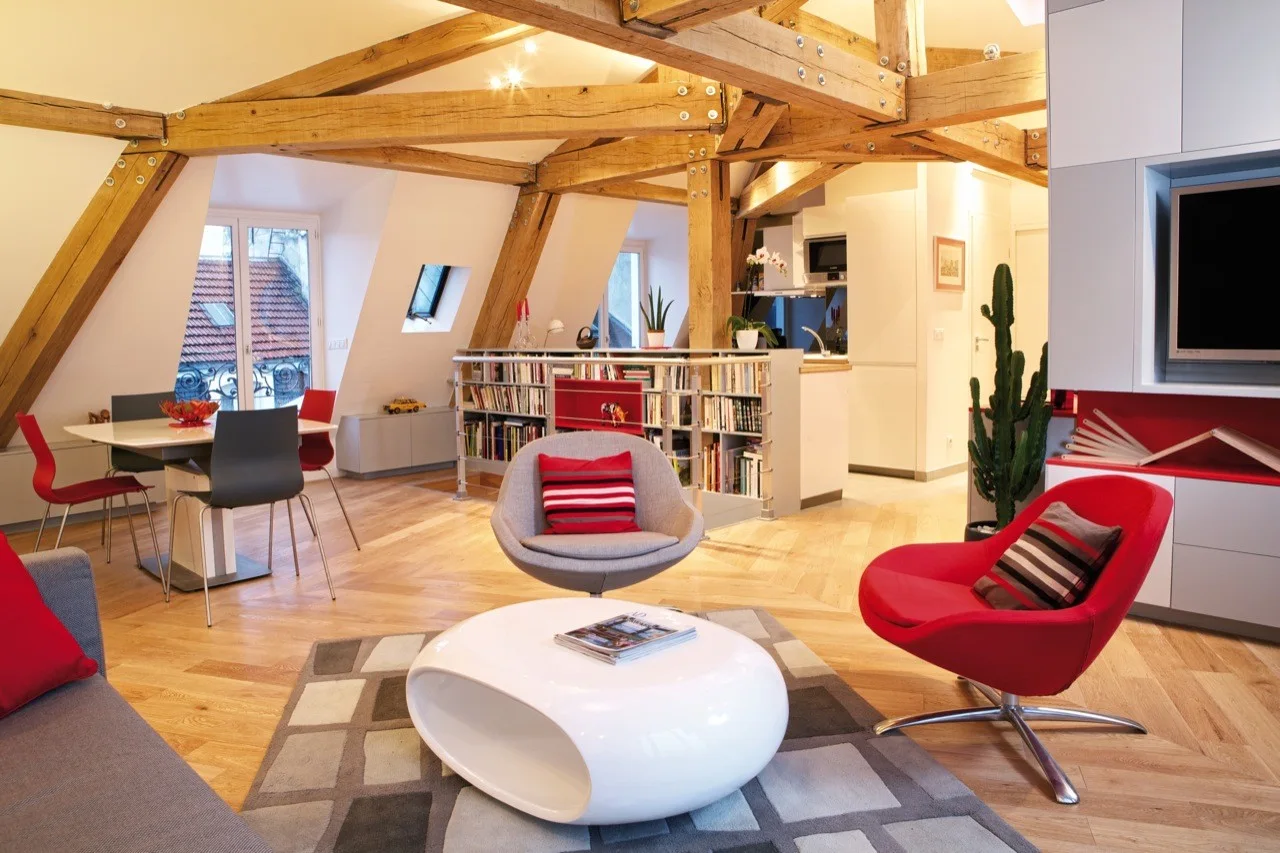 A modern loft living room features prominent exposed structural timber beams, light parquet flooring, red accent armchairs, and a low white coffee table.