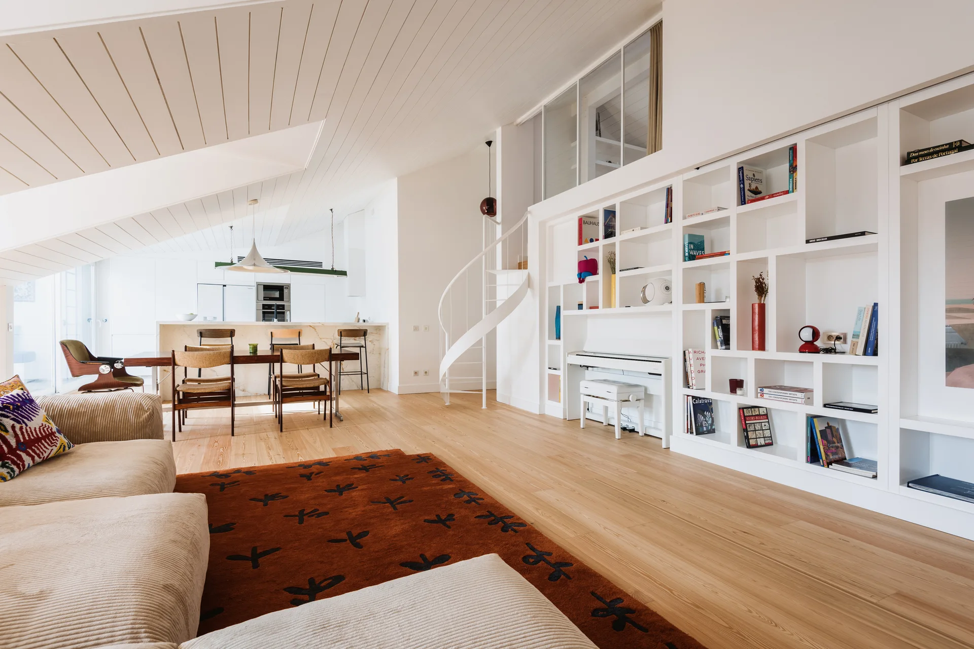 A modern open-plan living room features a beige sofa on a rust-colored rug, a white spiral staircase, and a large custom white built-in bookshelf.