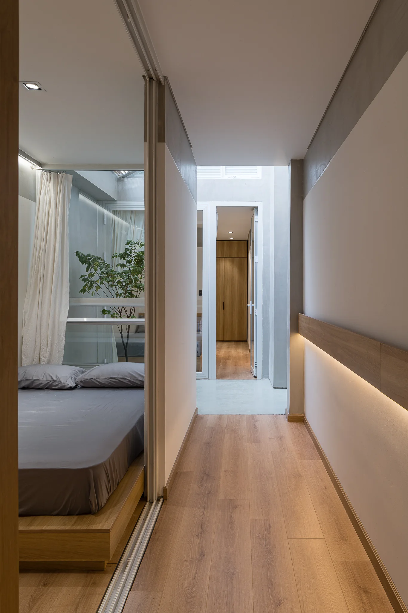 A view down a modern residential hallway with light wood floors and linear wall lighting next to a bedroom with a low wooden bed.