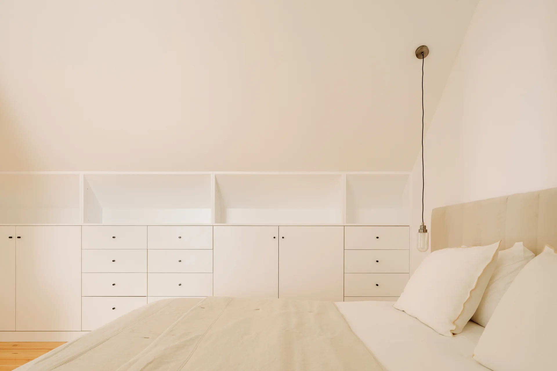 A minimalist loft bedroom with white built-in cabinetry and drawers beneath a sloped ceiling next to a bed with a beige headboard.