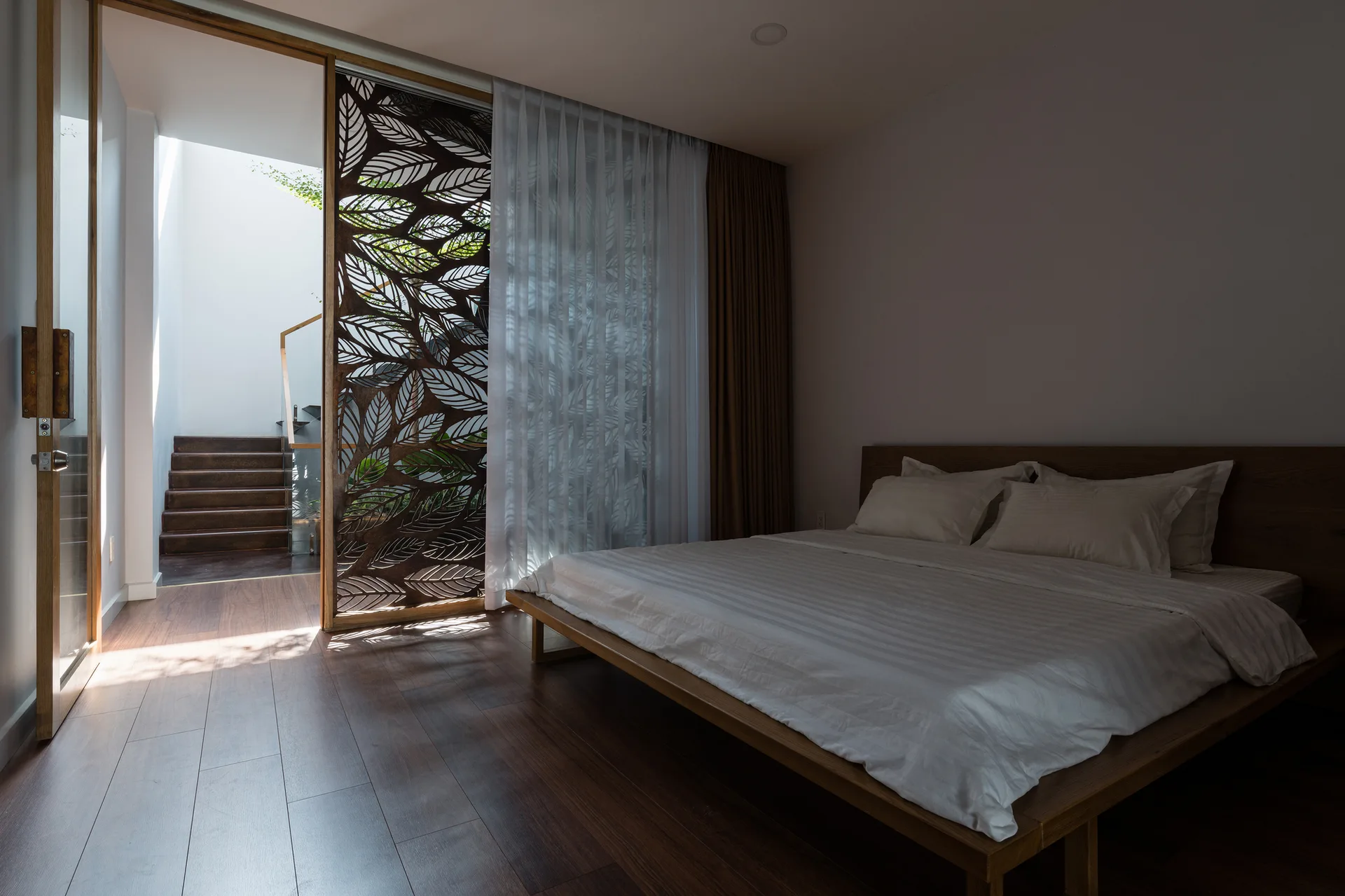 A modern bedroom features a wooden platform bed with white sheets next to a laser-cut decorative leaf screen showing a hallway with stairs.
