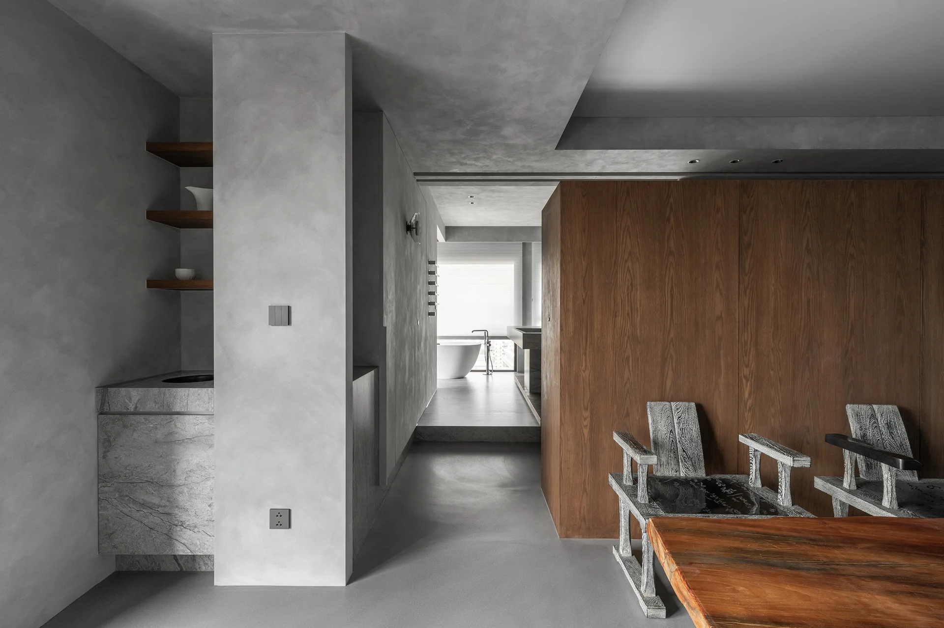 A minimalist concrete interior with a large wood paneled wall partition, sculptural chairs, and a corridor leading to a bright bathroom with a white tub.