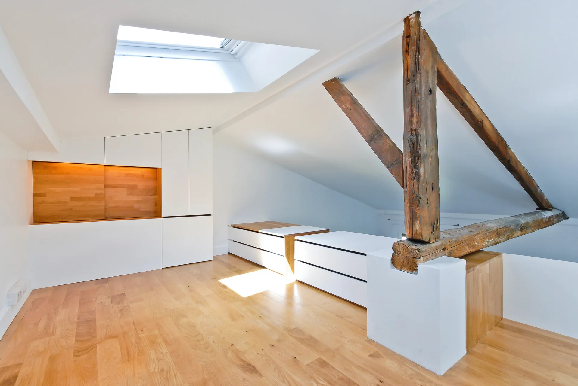 A minimalist attic bedroom featuring a skylight in the sloped ceiling, light wood flooring, an exposed wood pillar, and built-in white cabinets.