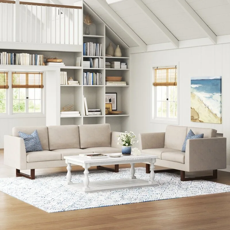 A light-filled living room features two beige sofas, a distressed white coffee table, and tall custom built-in bookshelves under a sloped vaulted ceiling.