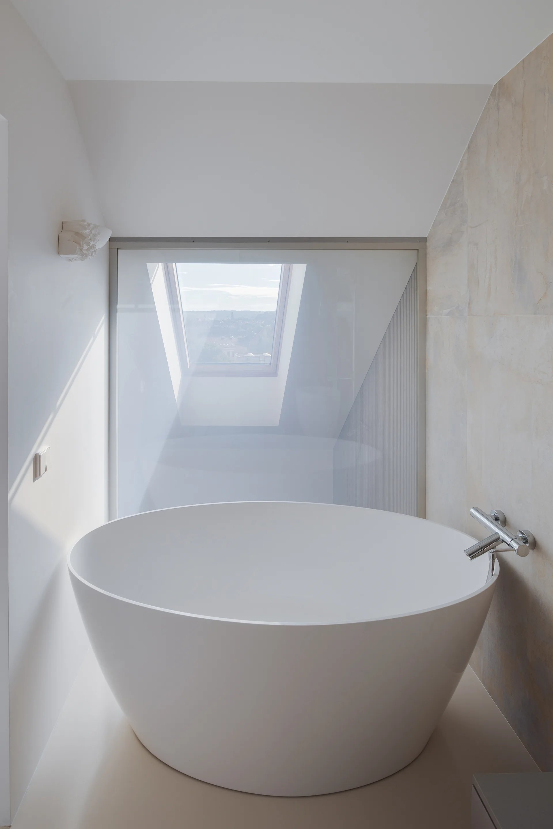 A circular white freestanding bathtub sits in an attic bathroom featuring a sloped ceiling, a large skylight, and a glass partition wall.