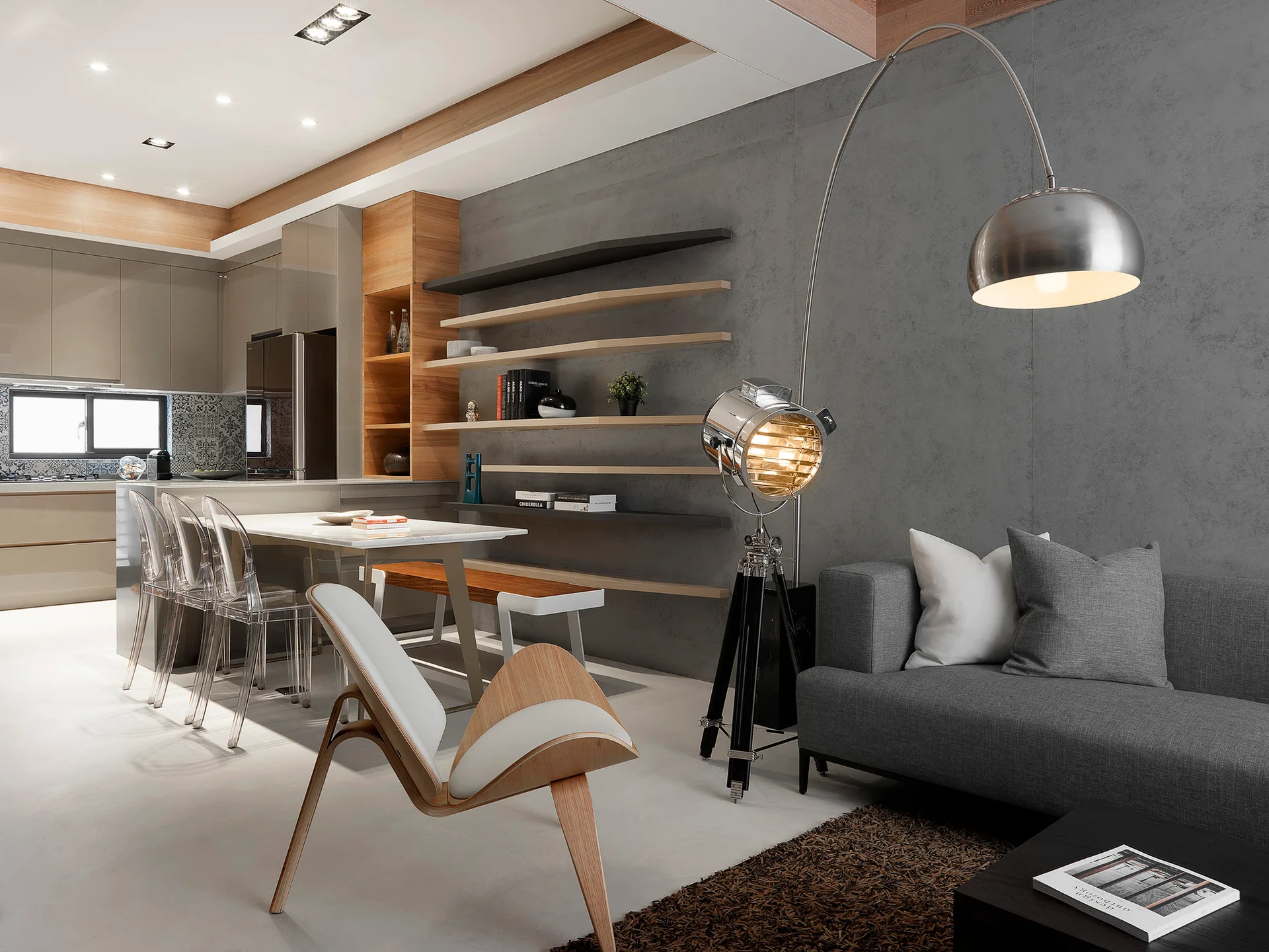 An open-plan living room features a grey sofa, a concrete-textured feature wall with floating wood shelves, and transparent acrylic dining chairs.